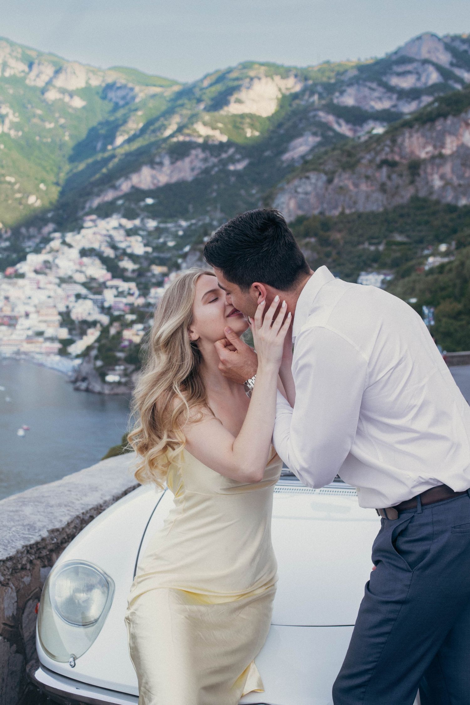 Couple sharing a kiss leaning against vintage white convertible with Positano cliffside village in background.
