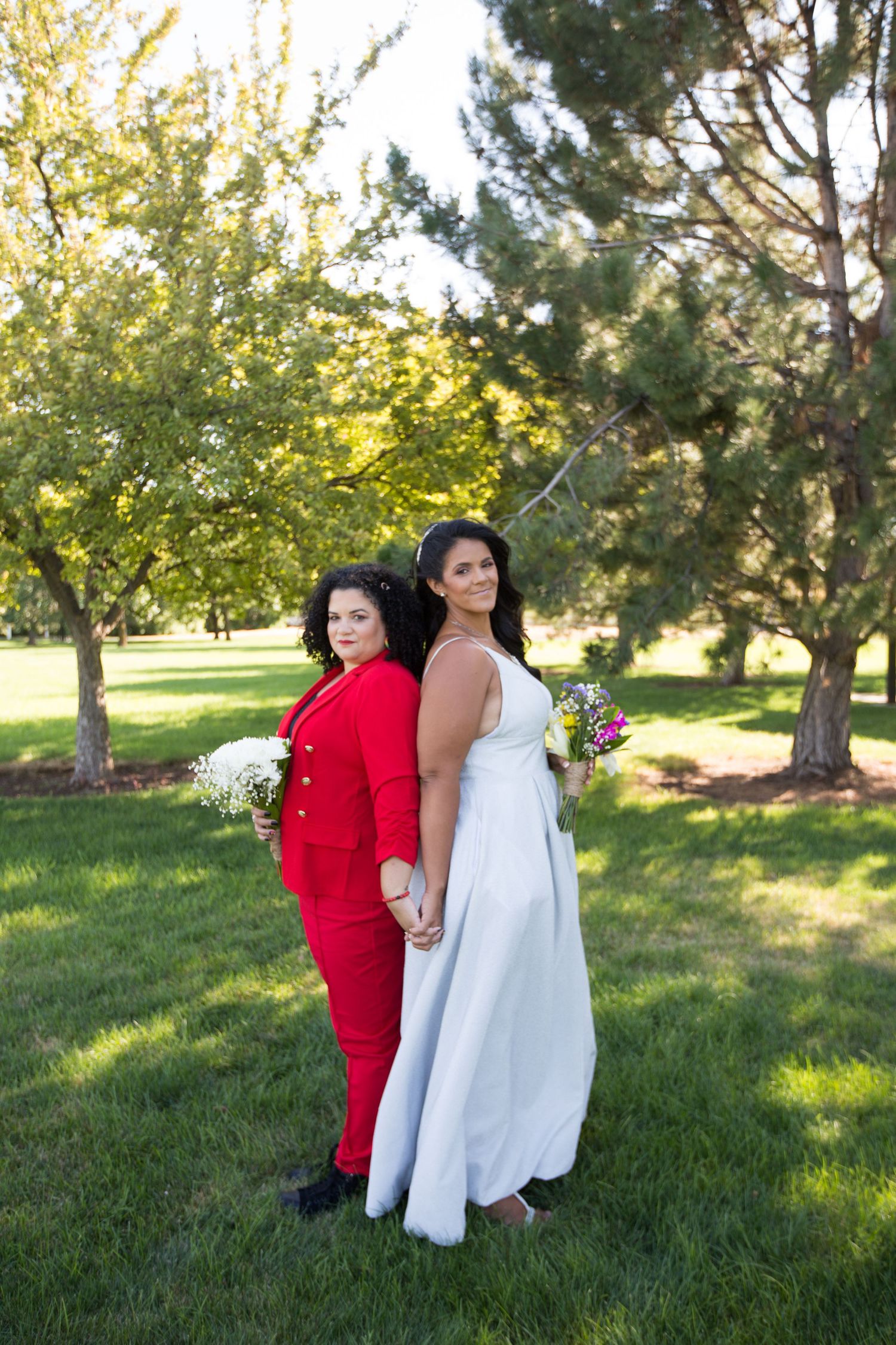 A couple in white and red attire poses together on grassy grounds with trees in the background.