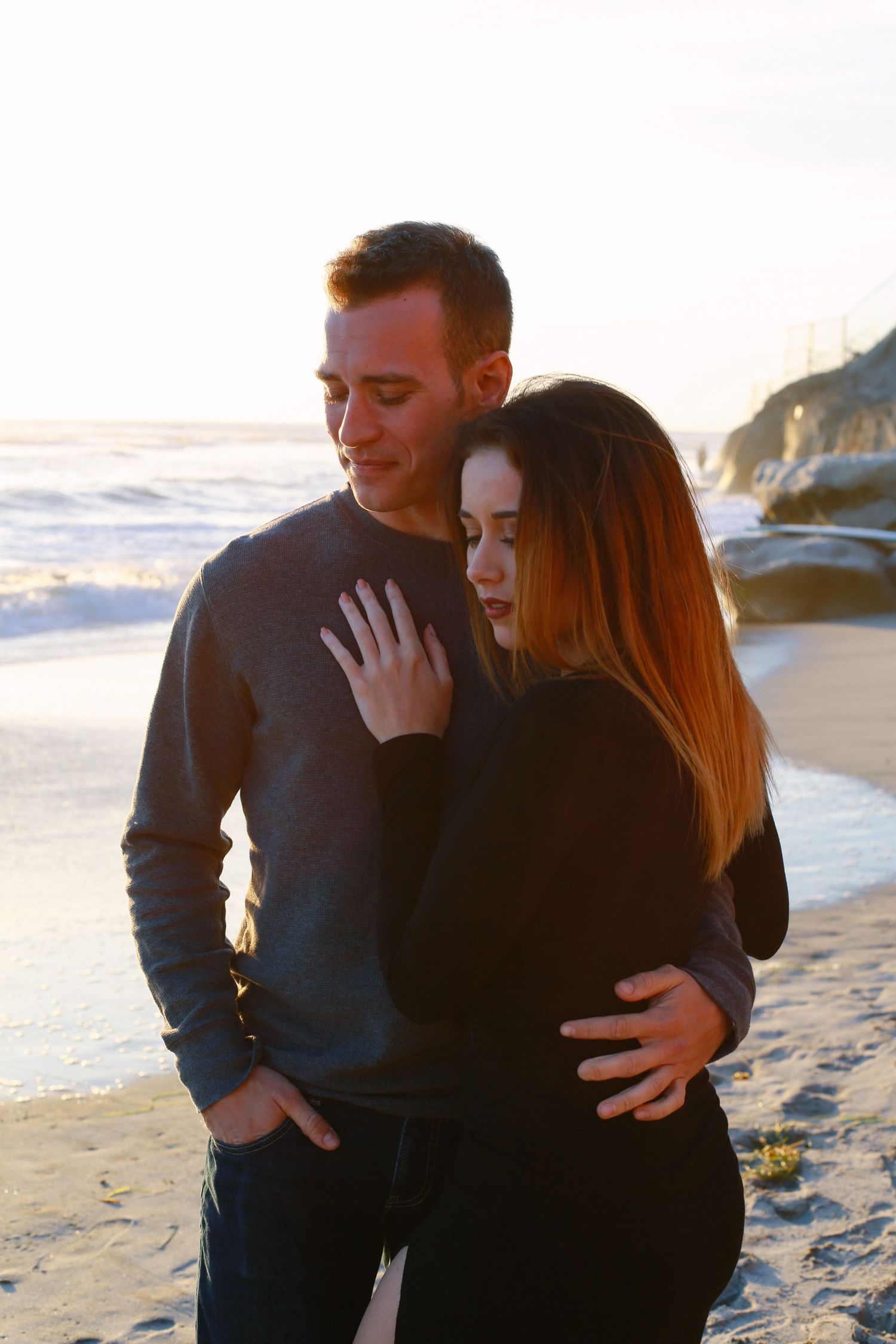 A couple in dark sweaters embraces on a beach at sunset with ocean waves in the background.