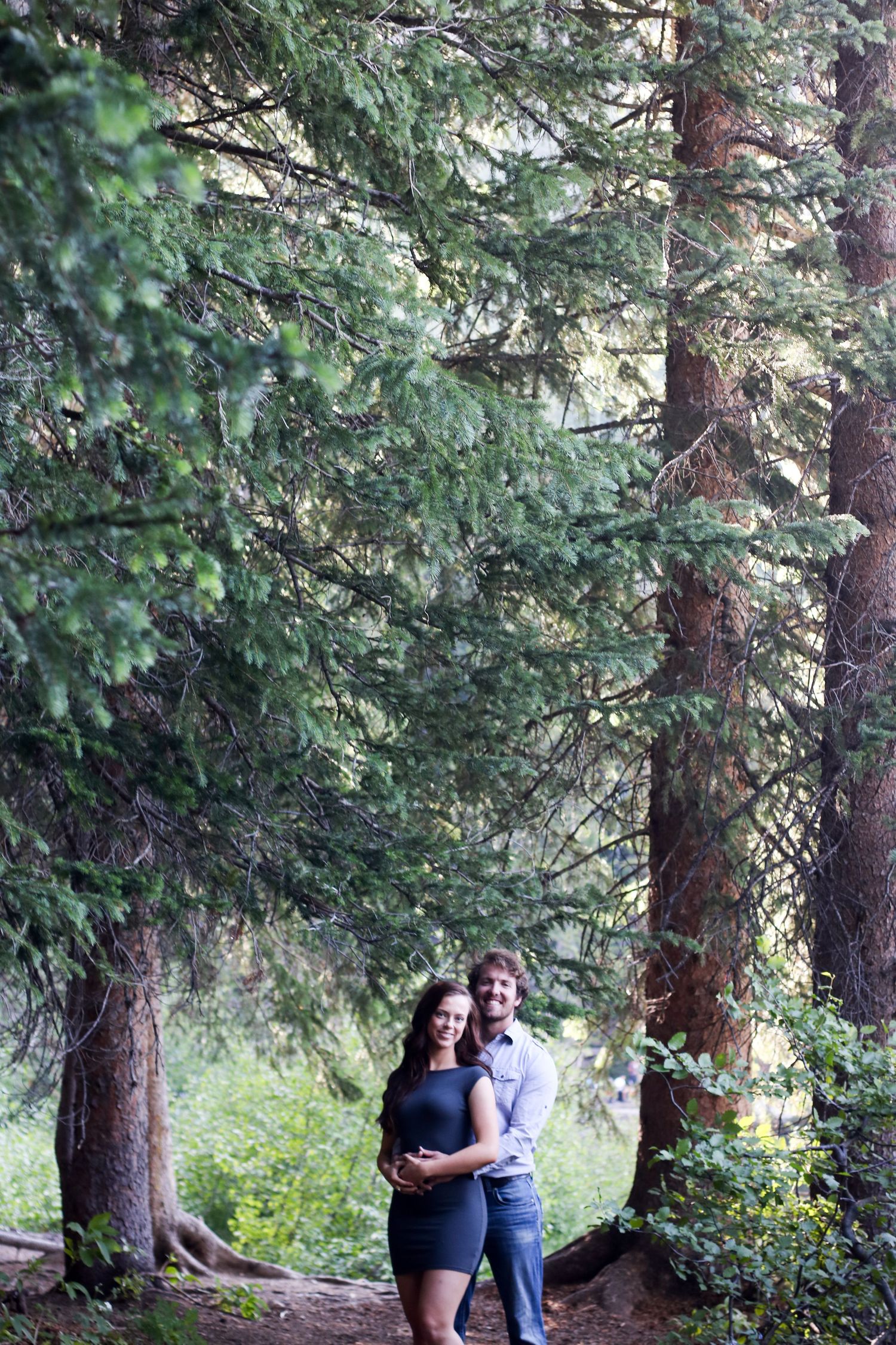 A couple shares a tender moment under tall evergreen trees in a forest setting.