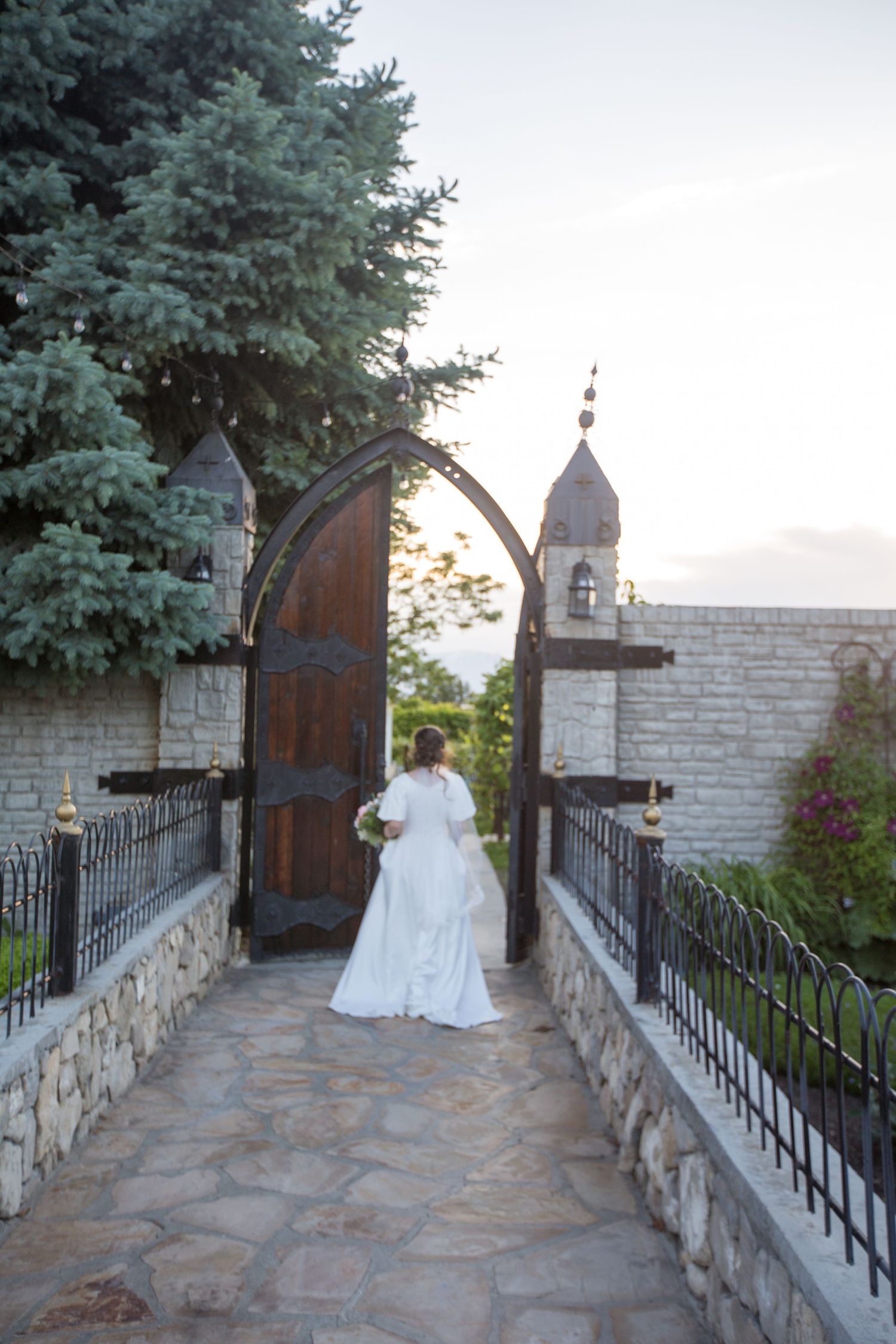 A stone pathway with wrought iron railings leads to an arched wooden door surrounded by evergreen trees.