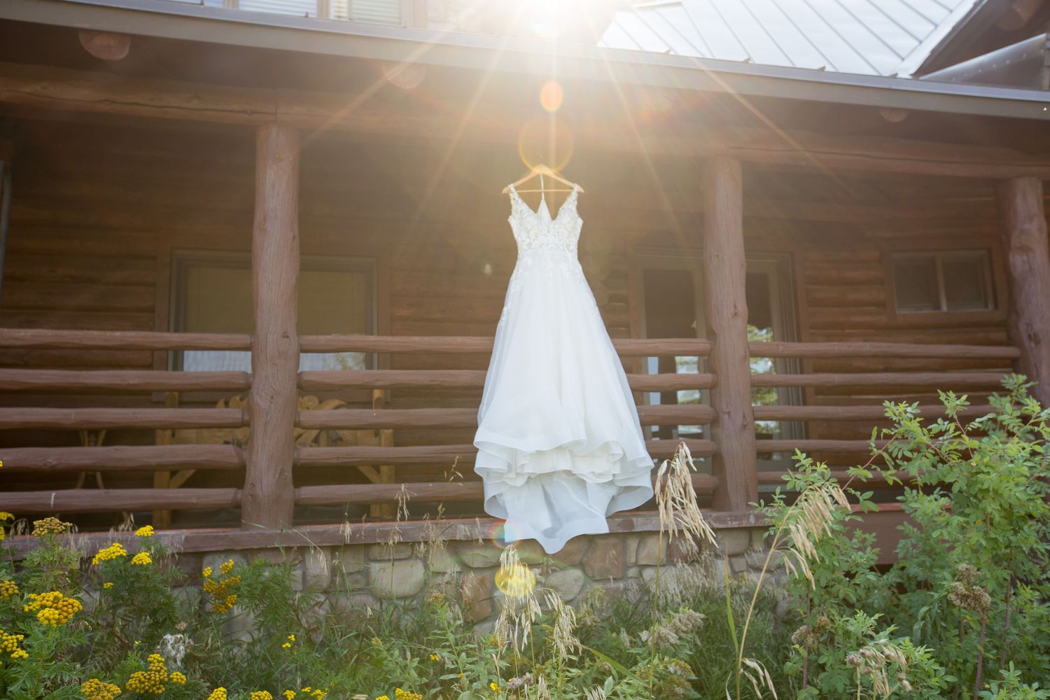 Wedding dress hanging outside rustic cabin with sunflare and wildflowers.