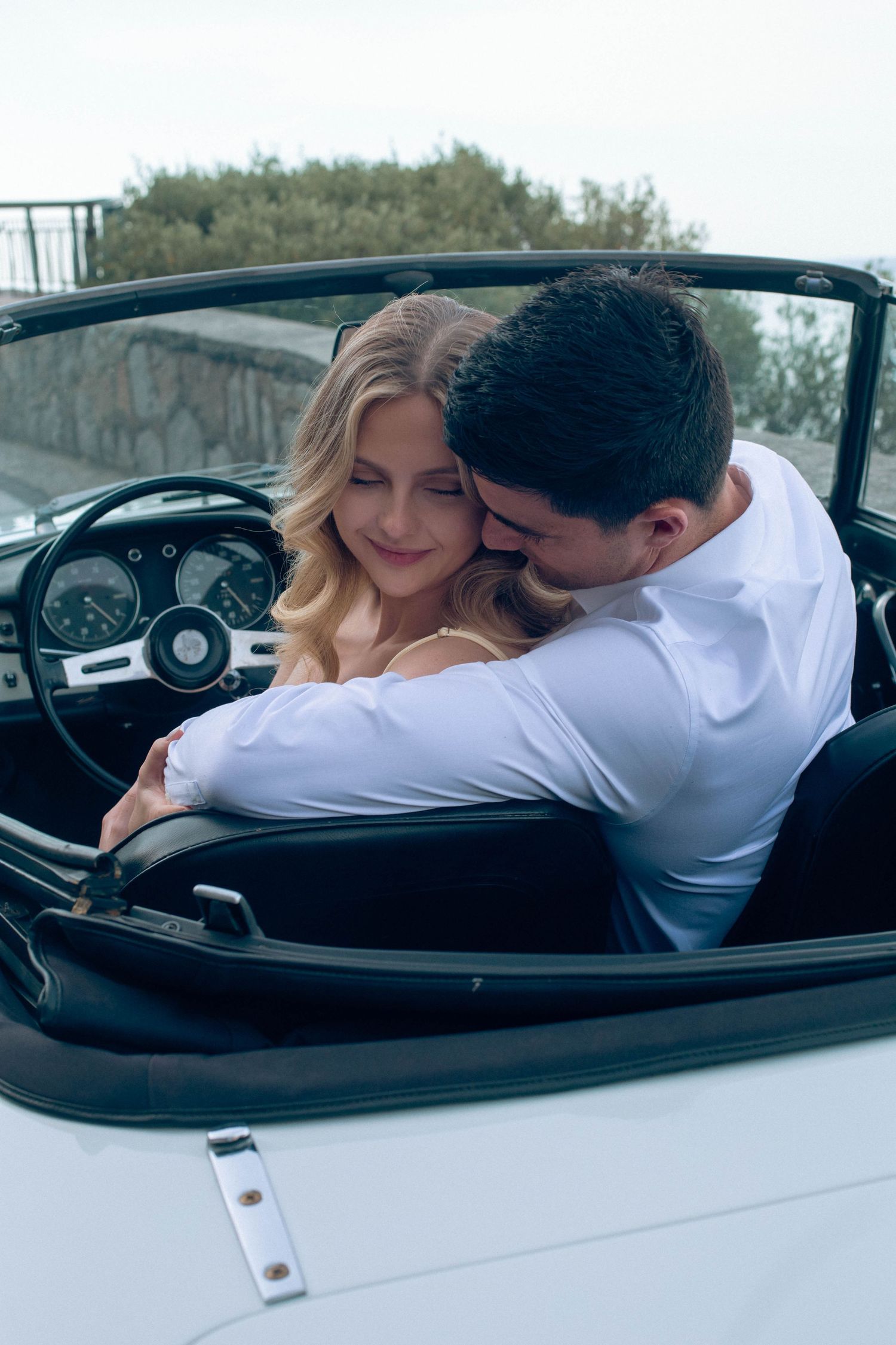 Romantic couple embracing in a classic white convertible car, woman in white dress smiling softly.