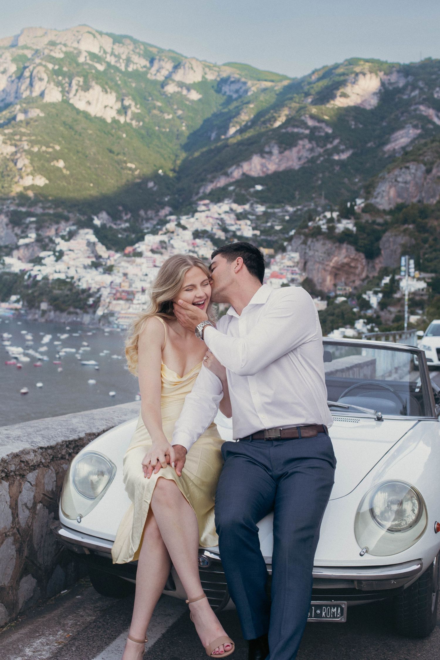 Couple laughing together leaning on vintage white convertible with Positano coastal village backdrop.