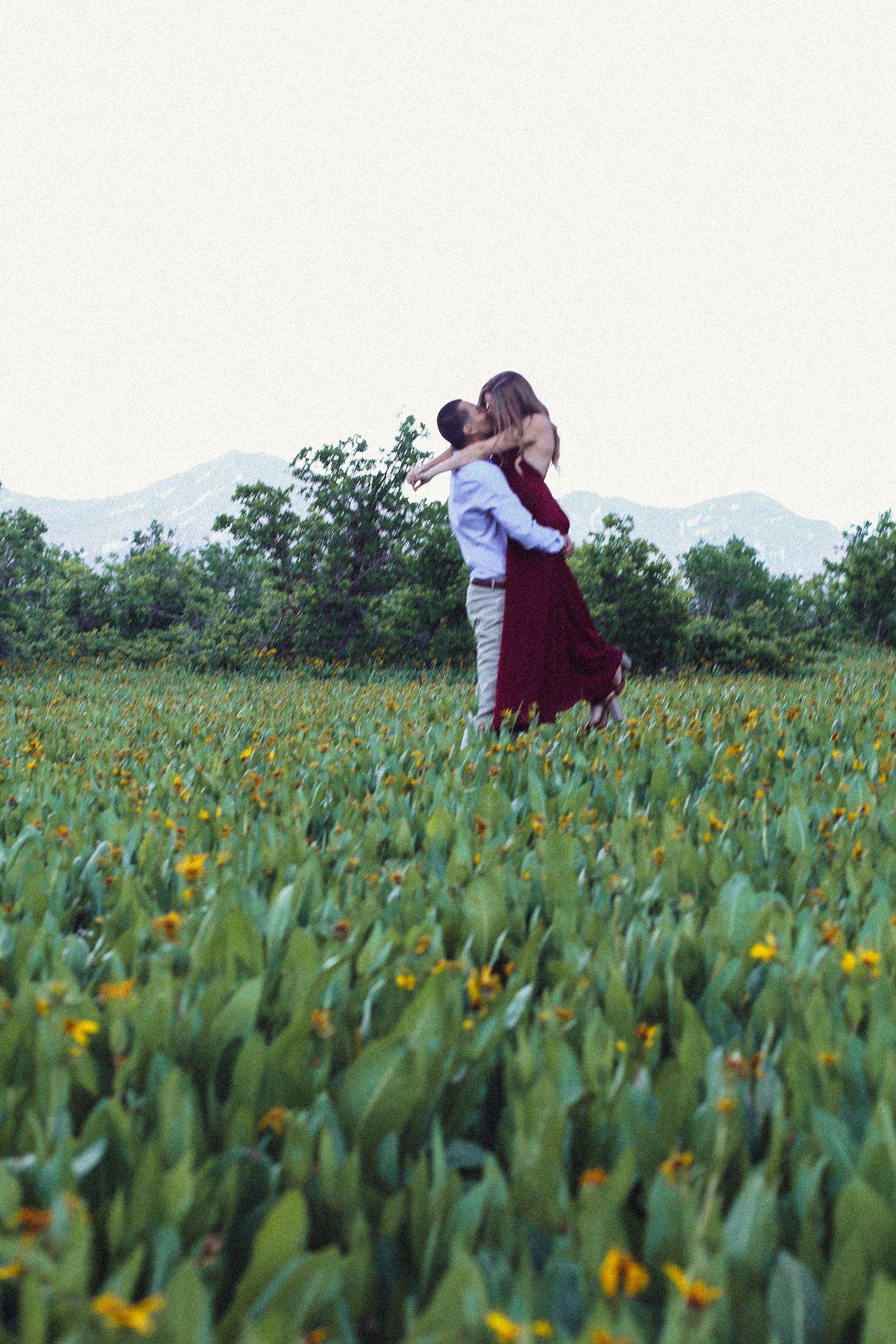 Couple embracing and kissing in a lush green field with yellow wildflowers and mountains in the background.