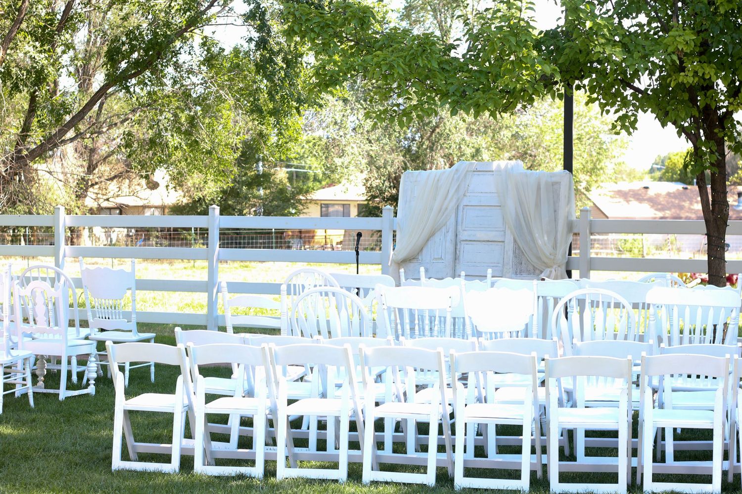 Outdoor wedding ceremony setup with rows of white chairs under green trees.