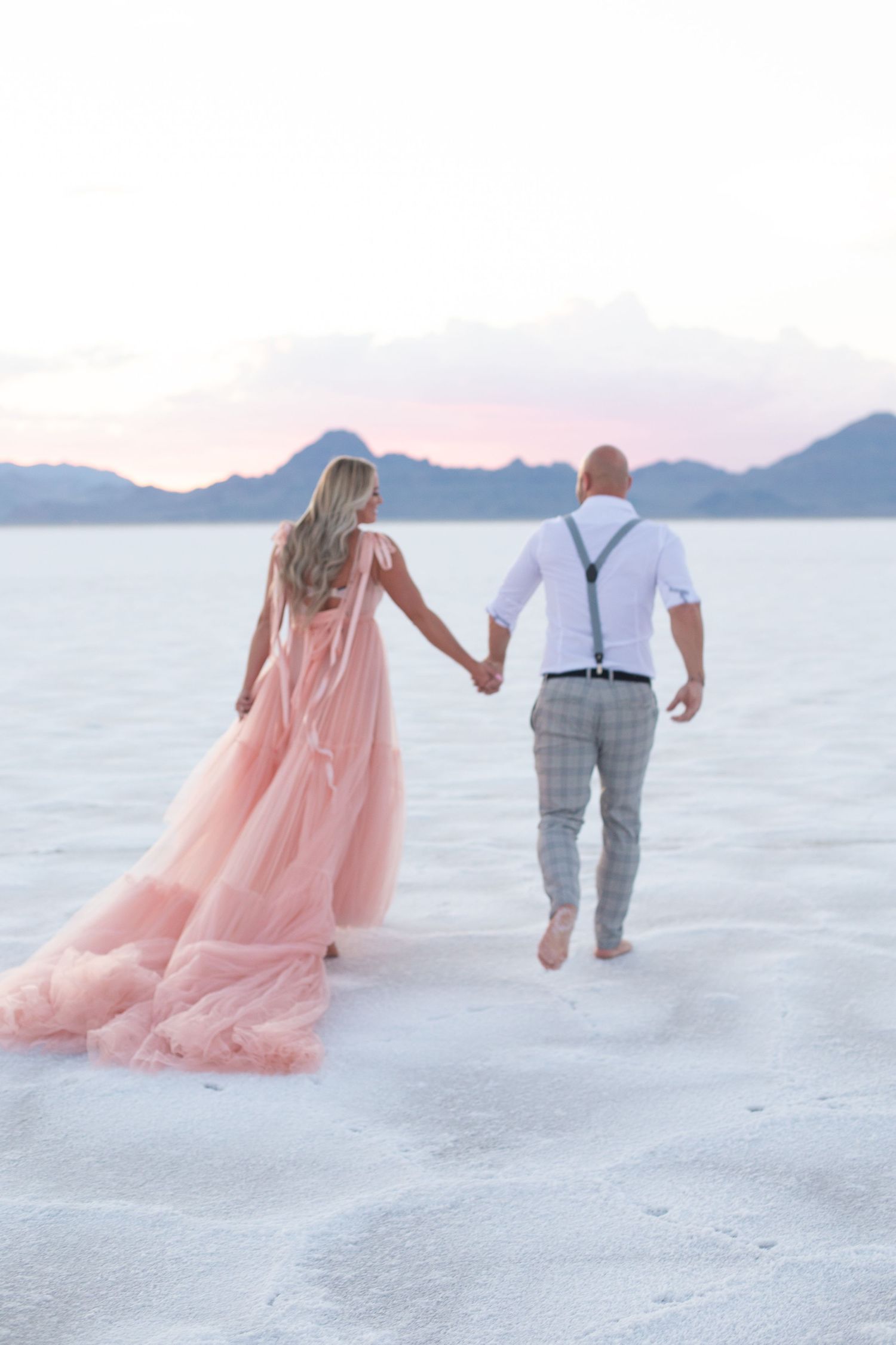 Couple walks away holding hands on salt flats with pink dress flowing in breeze.