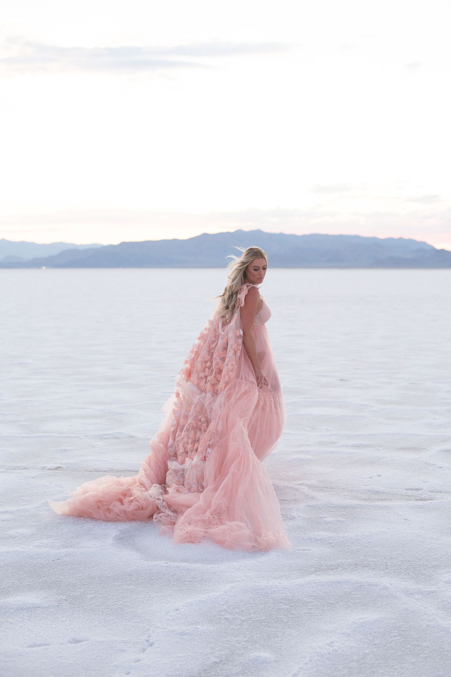 A flowing pink gown creates ethereal movement against a serene white landscape and mountain backdrop.