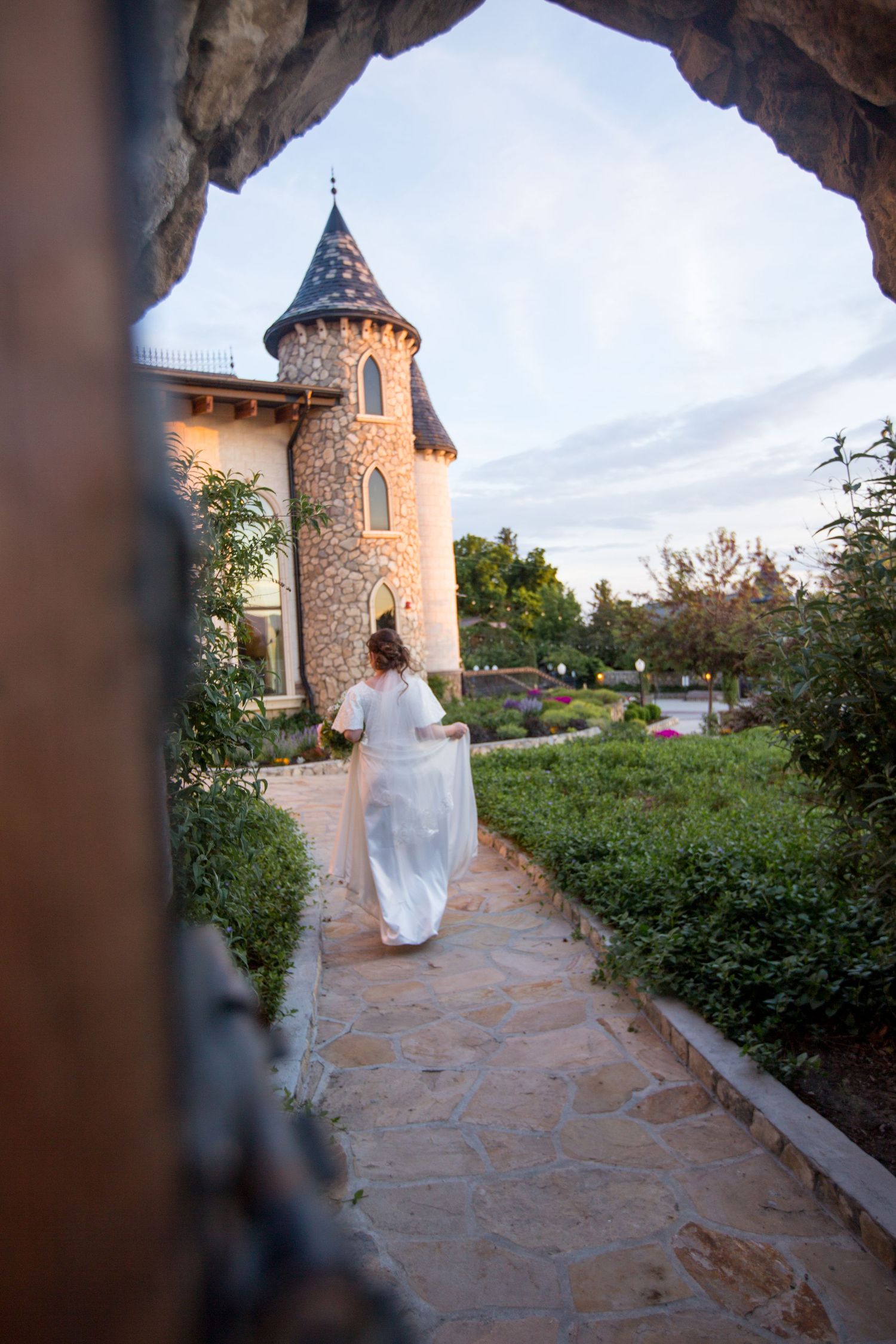 A bride in white walking along a stone pathway at a castle-style venue during sunset.