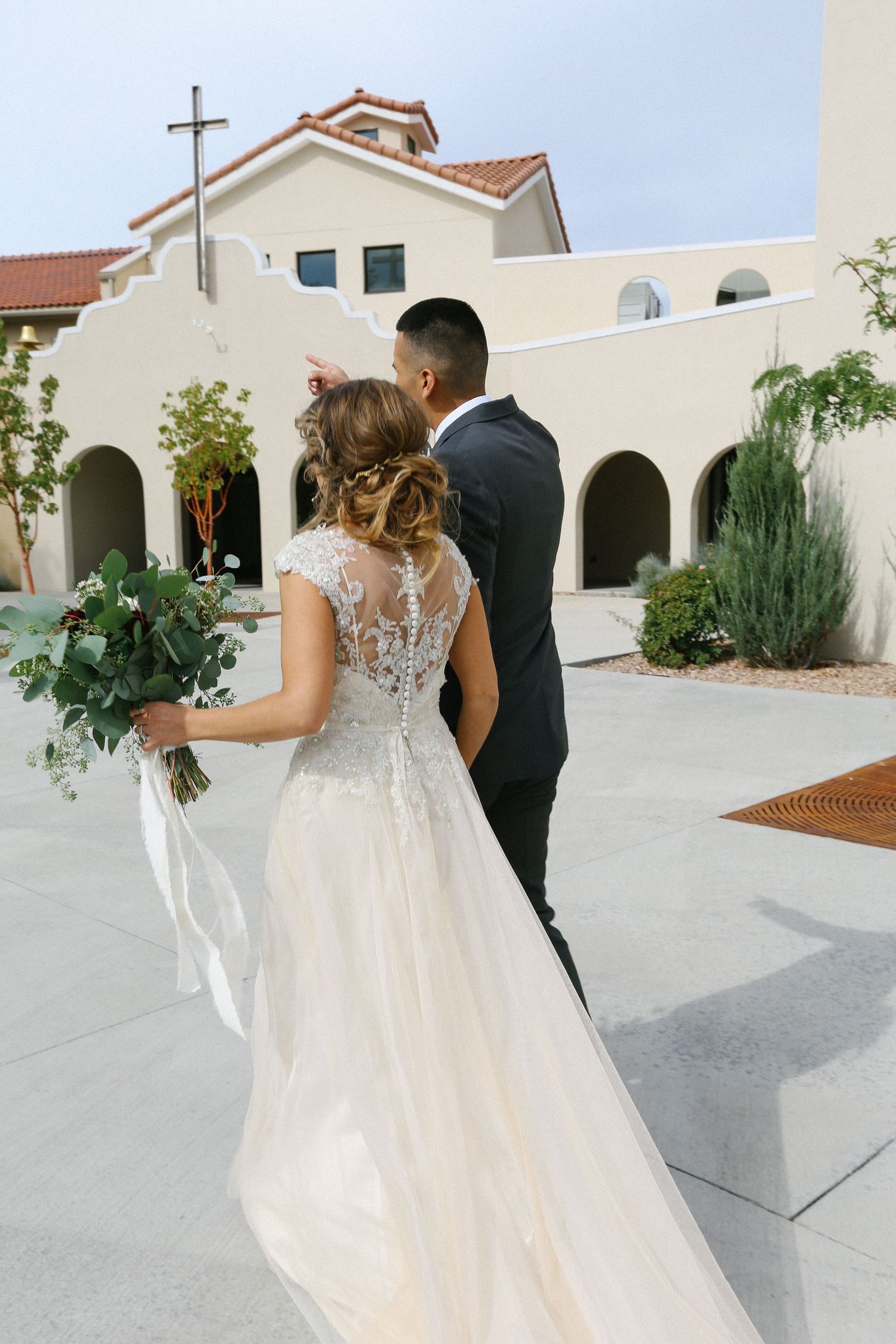 A bride and groom stand outside a Spanish-style church with white stucco walls and terracotta roof.