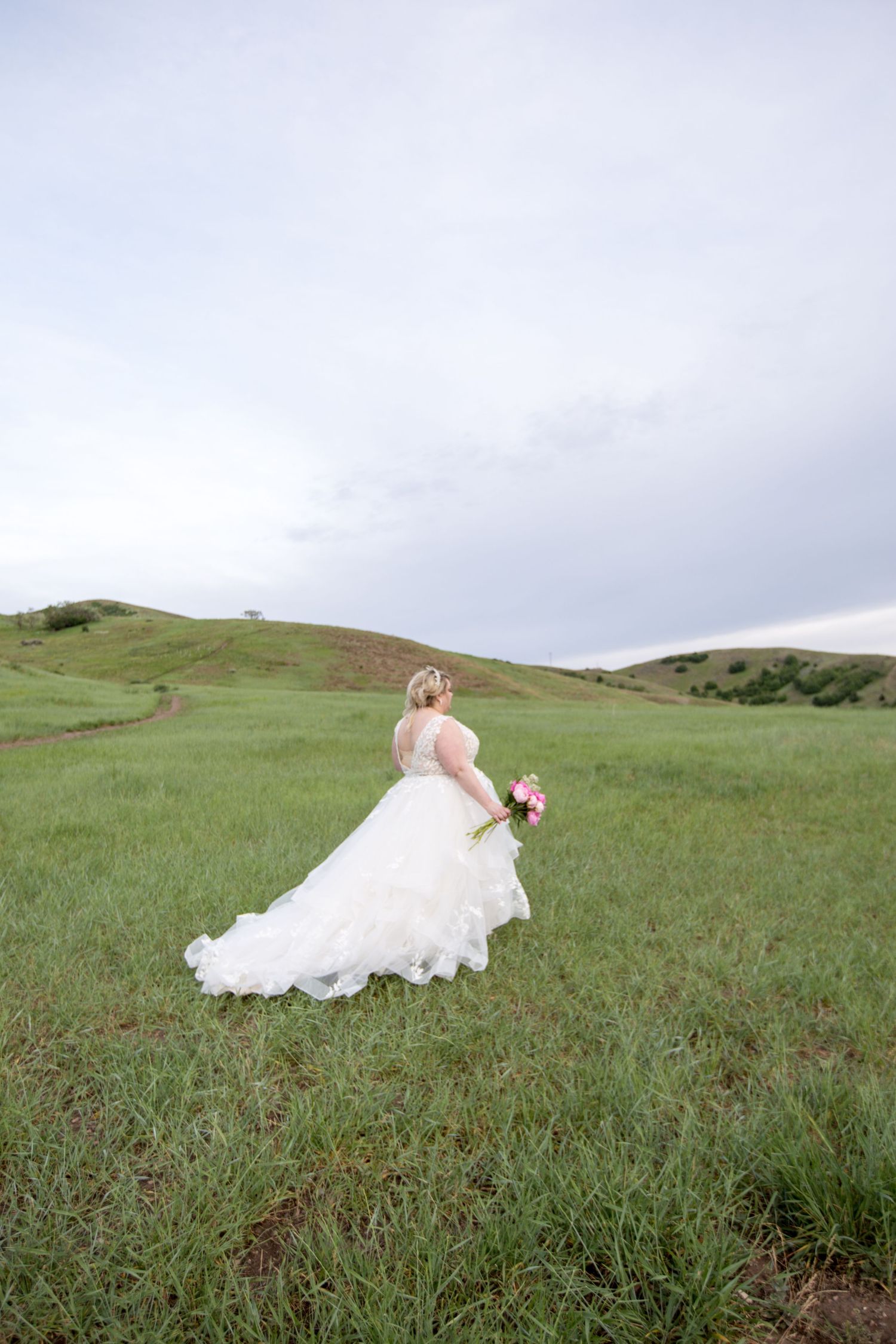 Bride in flowing white gown stands in green field holding bouquet.