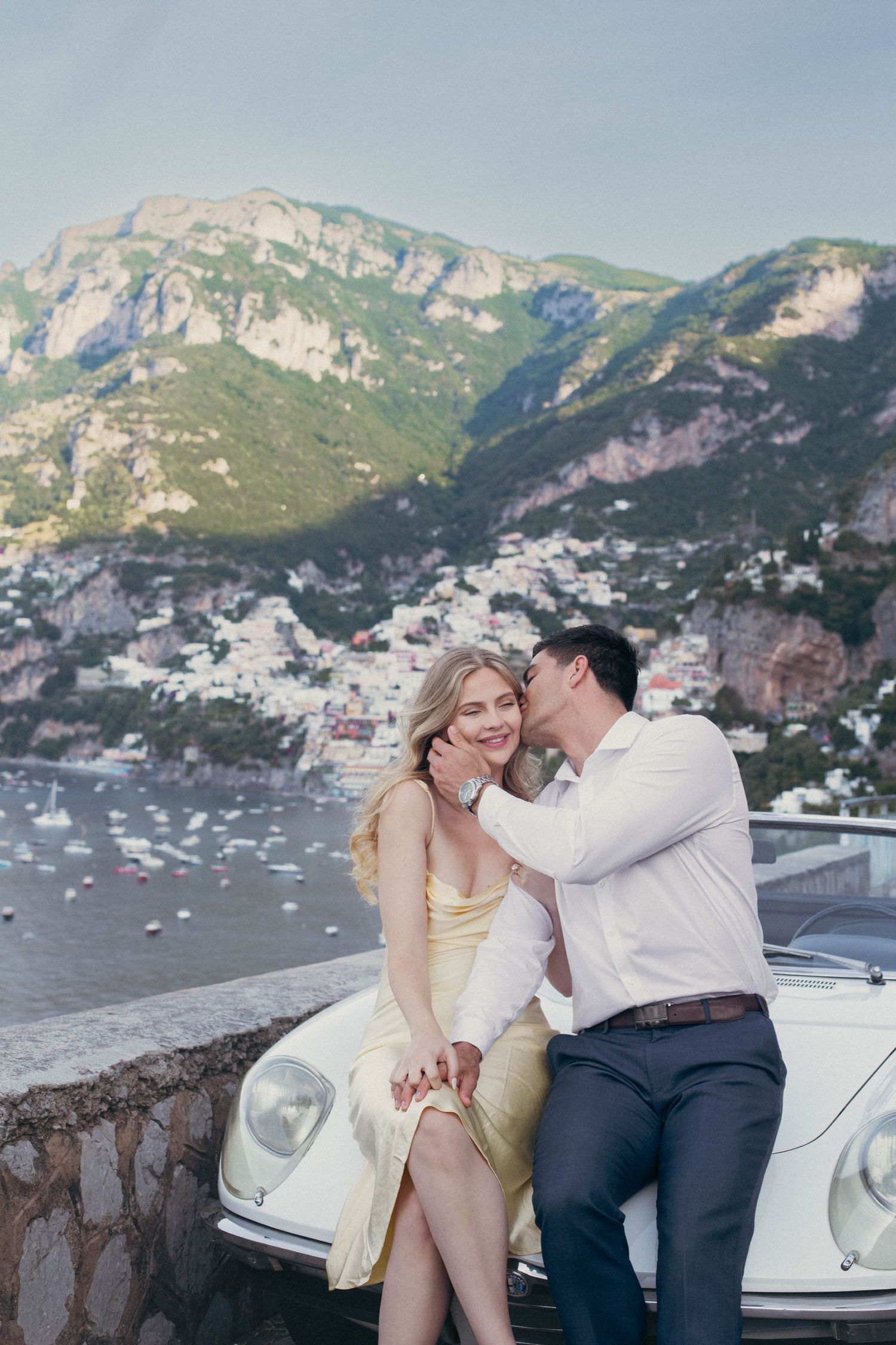 Romantic couple sharing intimate moment leaning against vintage white convertible on Amalfi Coast overlook.