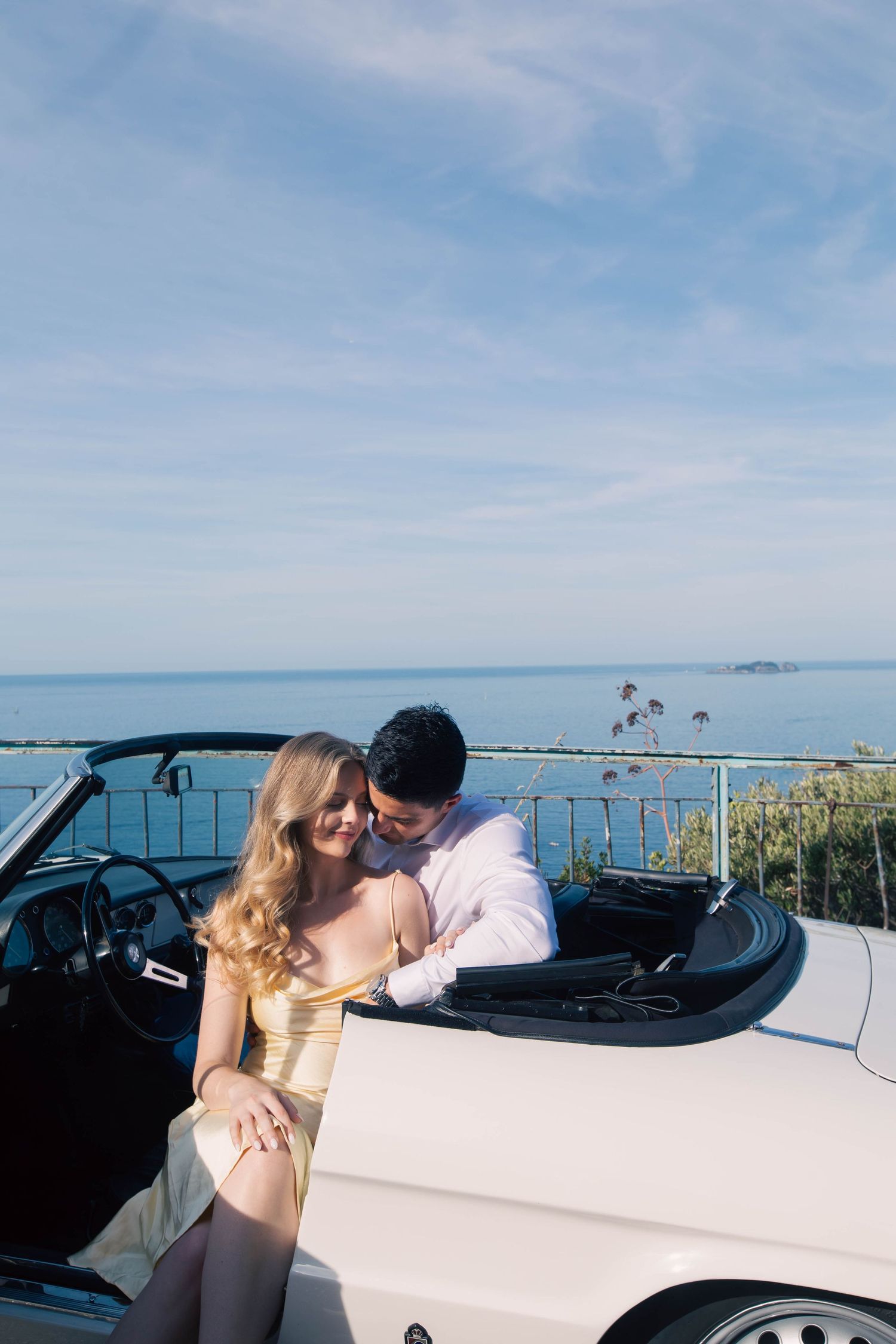 Couple embracing beside a vintage white convertible with panoramic Mediterranean Sea views on the Amalfi Coast.