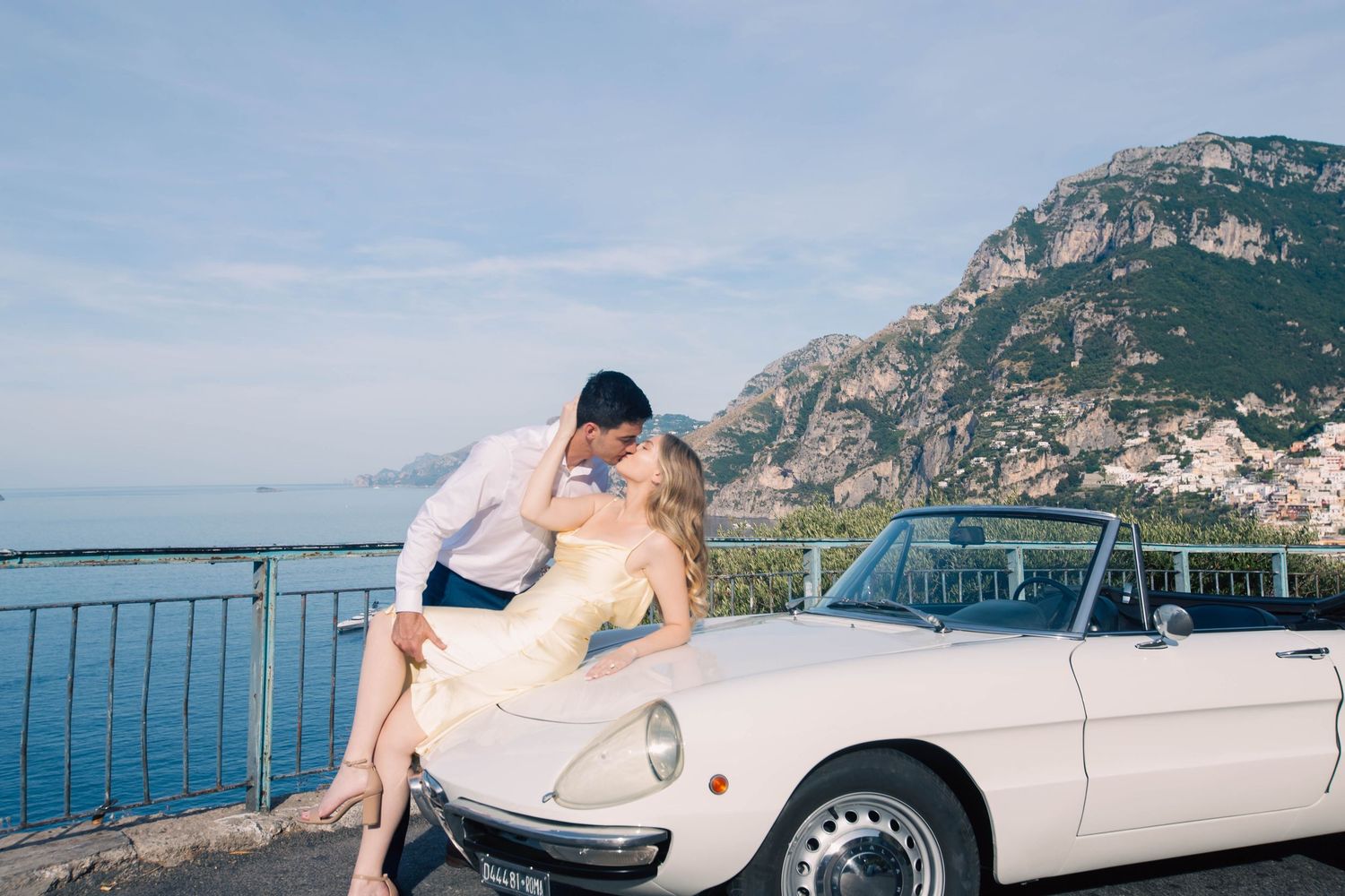 Couple kissing passionately beside white vintage Alfa Romeo Spider with Positano and blue sea backdrop.