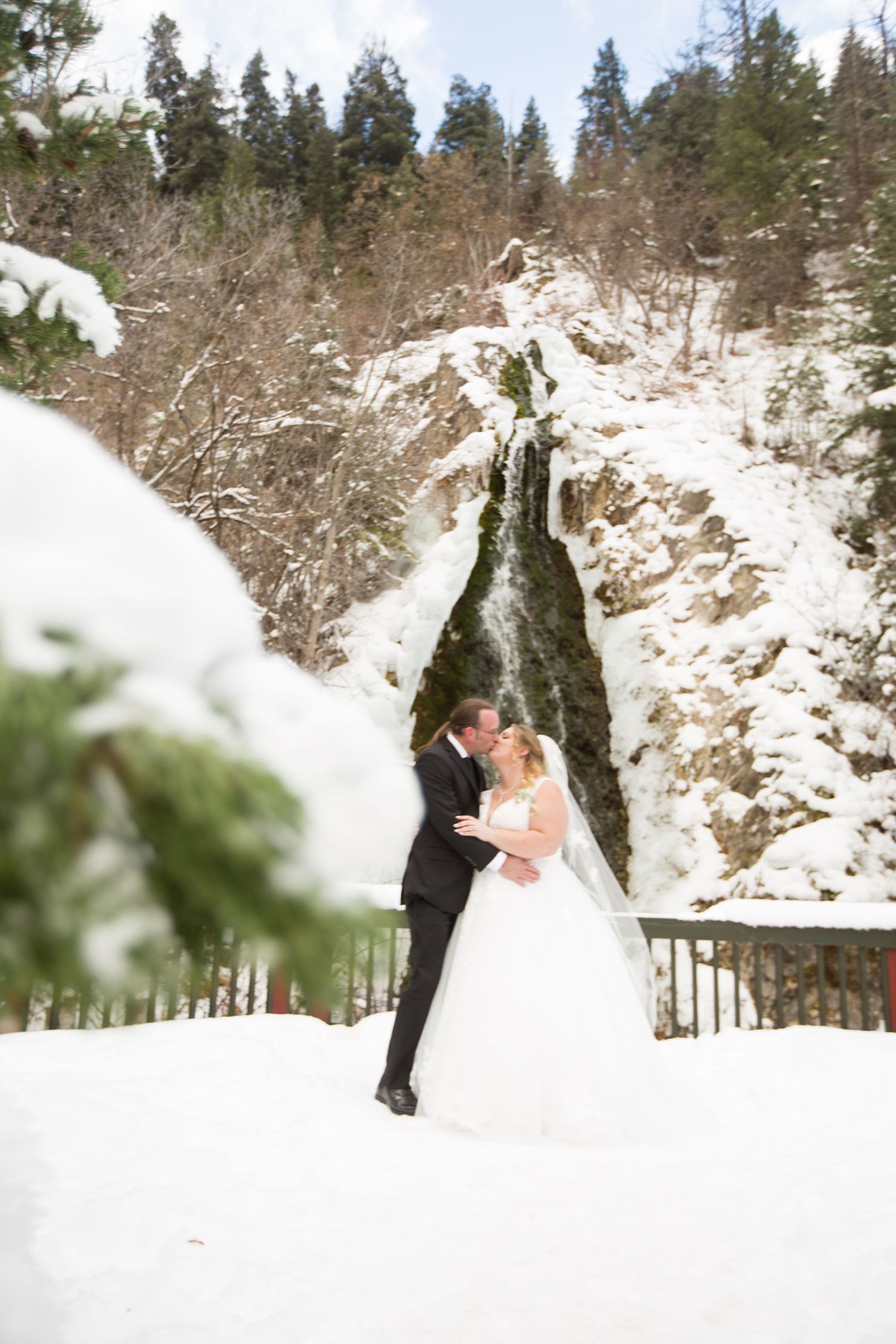 Wedding couple poses together in a snowy winter landscape with evergreen trees in the background.