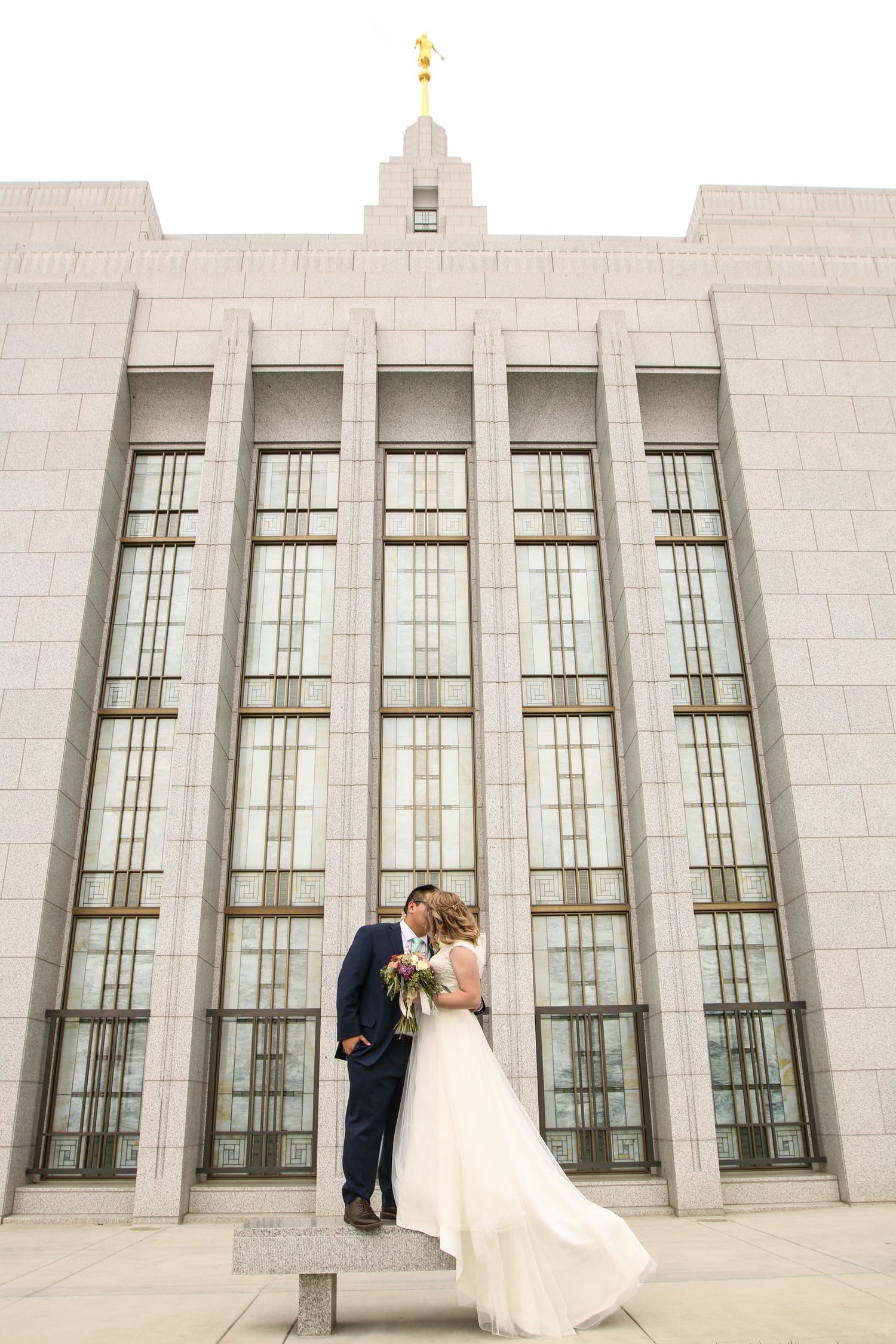 Wedding couple embracing in front of a modern white building with tall windows.