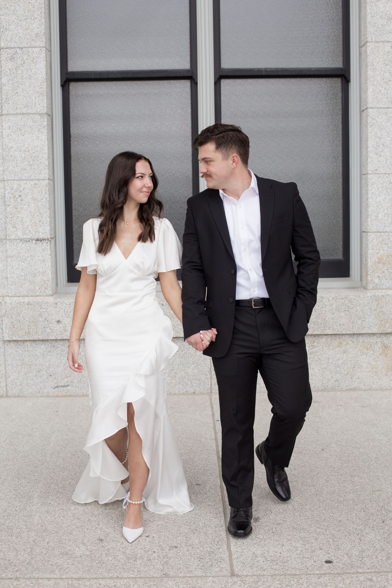 Wedding couple walks and poses along a modern building exterior with large windows.