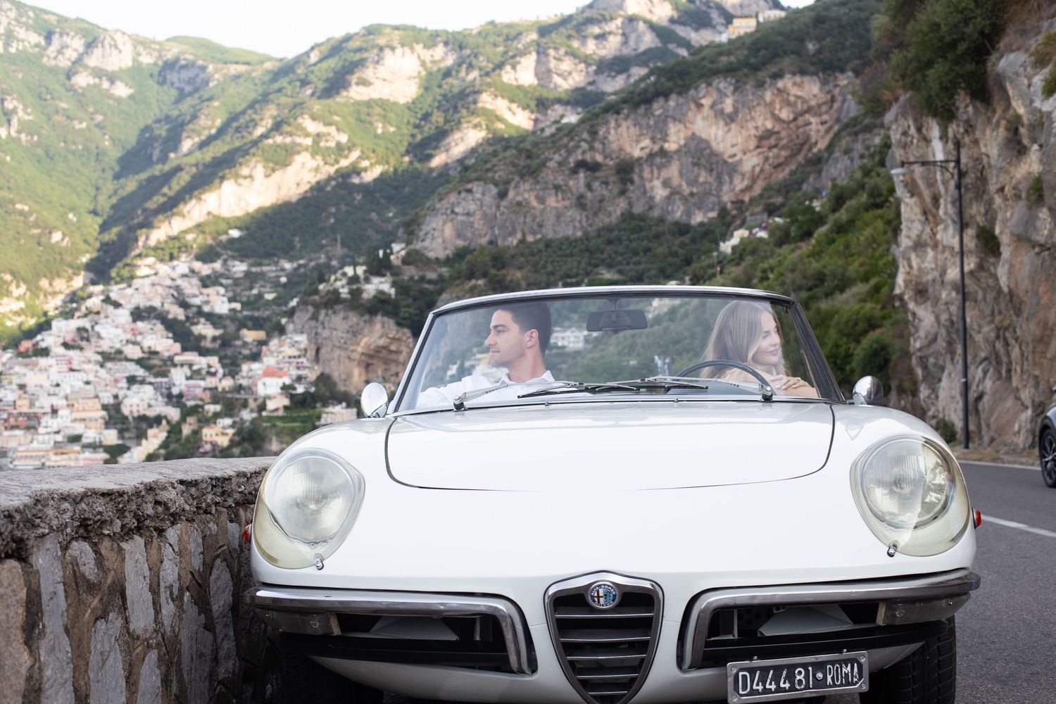 Couple driving a classic white Alfa Romeo convertible on a scenic Amalfi Coast road with mountains and village views.