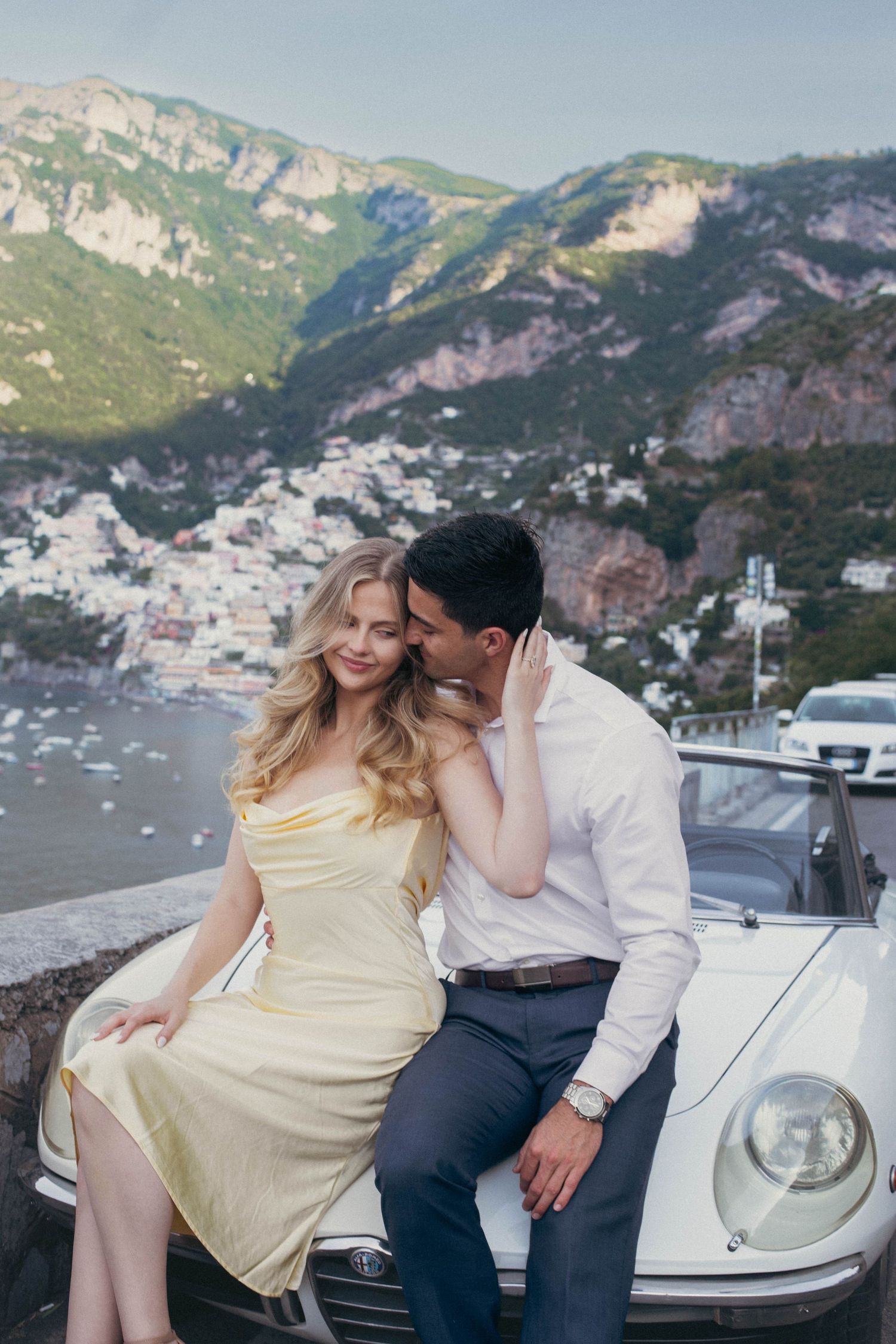 Couple sitting on vintage white car hood with Positano coastal village and mountains in background.
