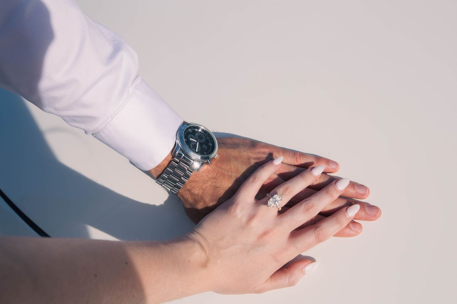 Close-up of couple's hands with engagement ring and luxury watch resting on a white car surface.
