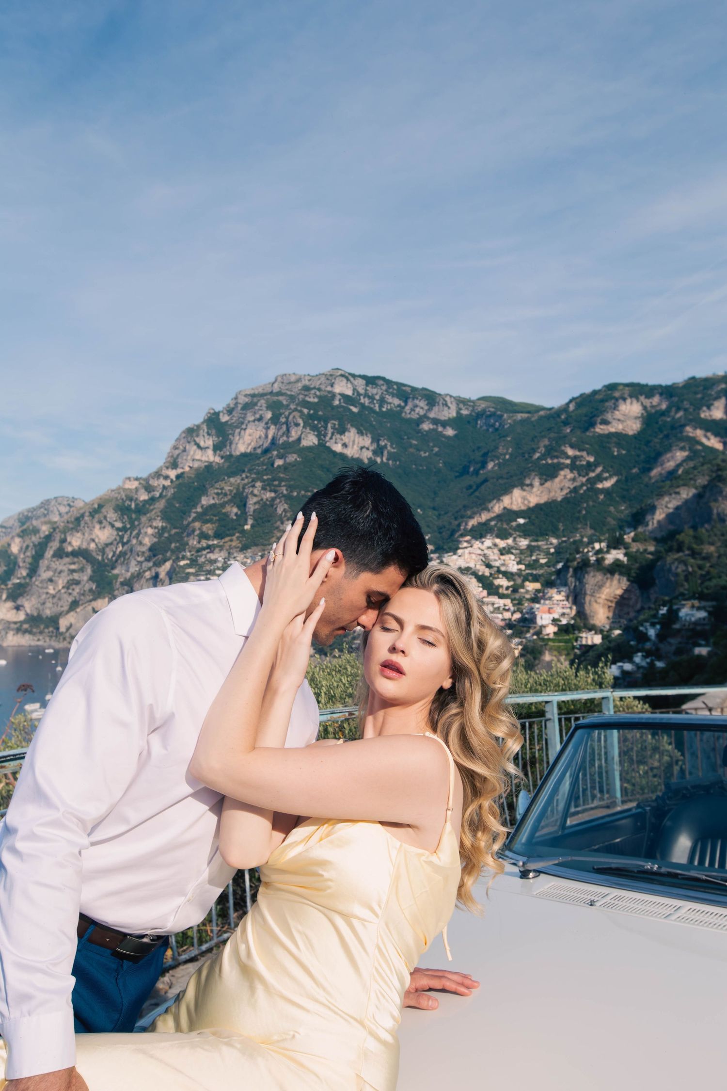 Romantic couple embracing by a white convertible with stunning Amalfi Coast mountains in the background.