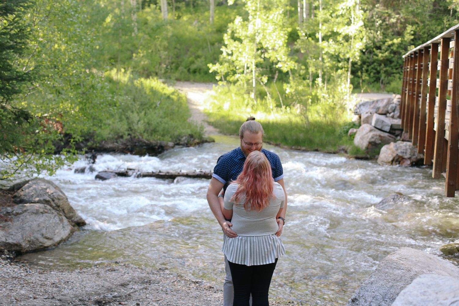 Couple embracing near a rushing mountain stream surrounded by lush green trees and rocky landscape.