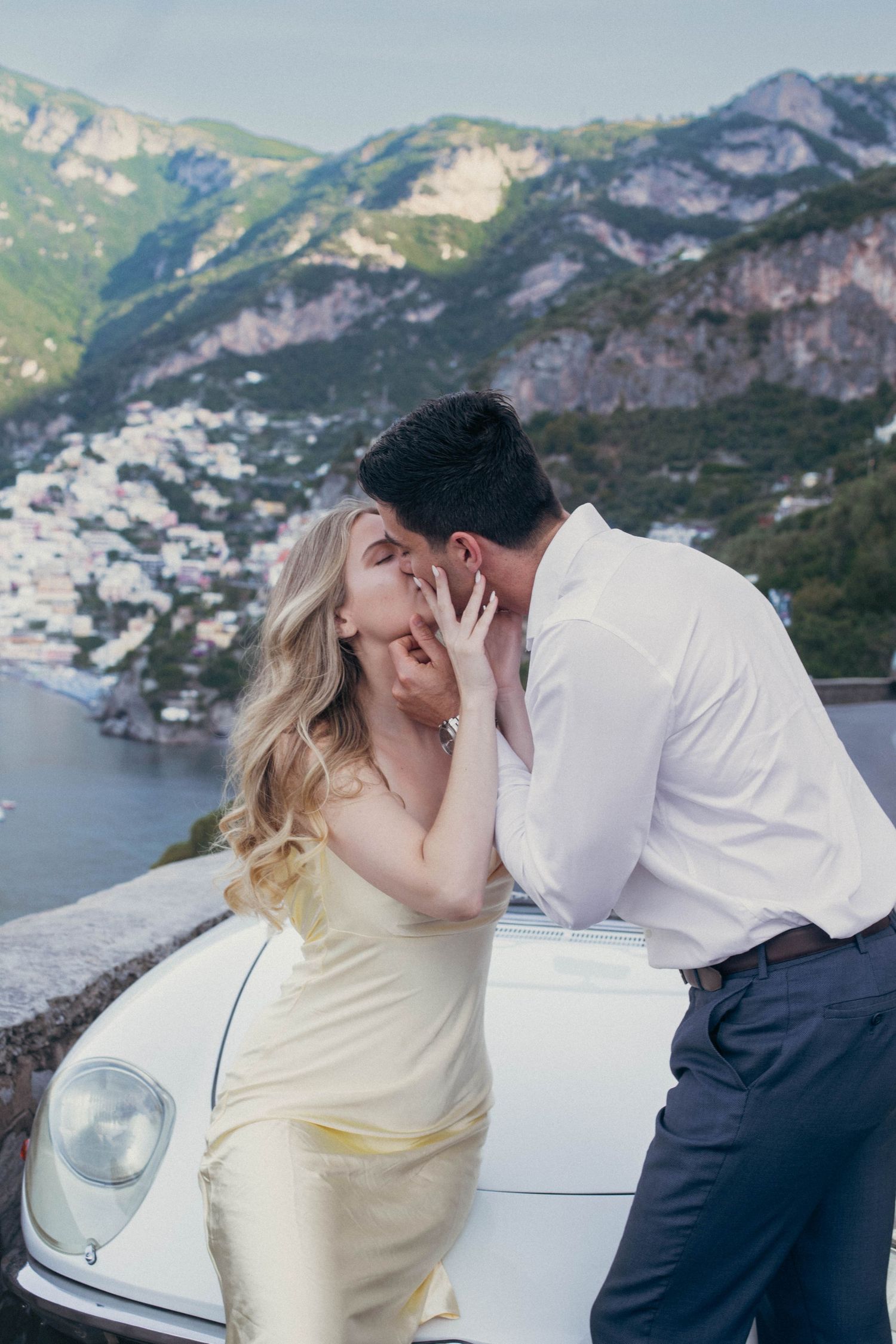 Romantic couple kissing beside a classic white convertible car with stunning Amalfi Coast mountain views.