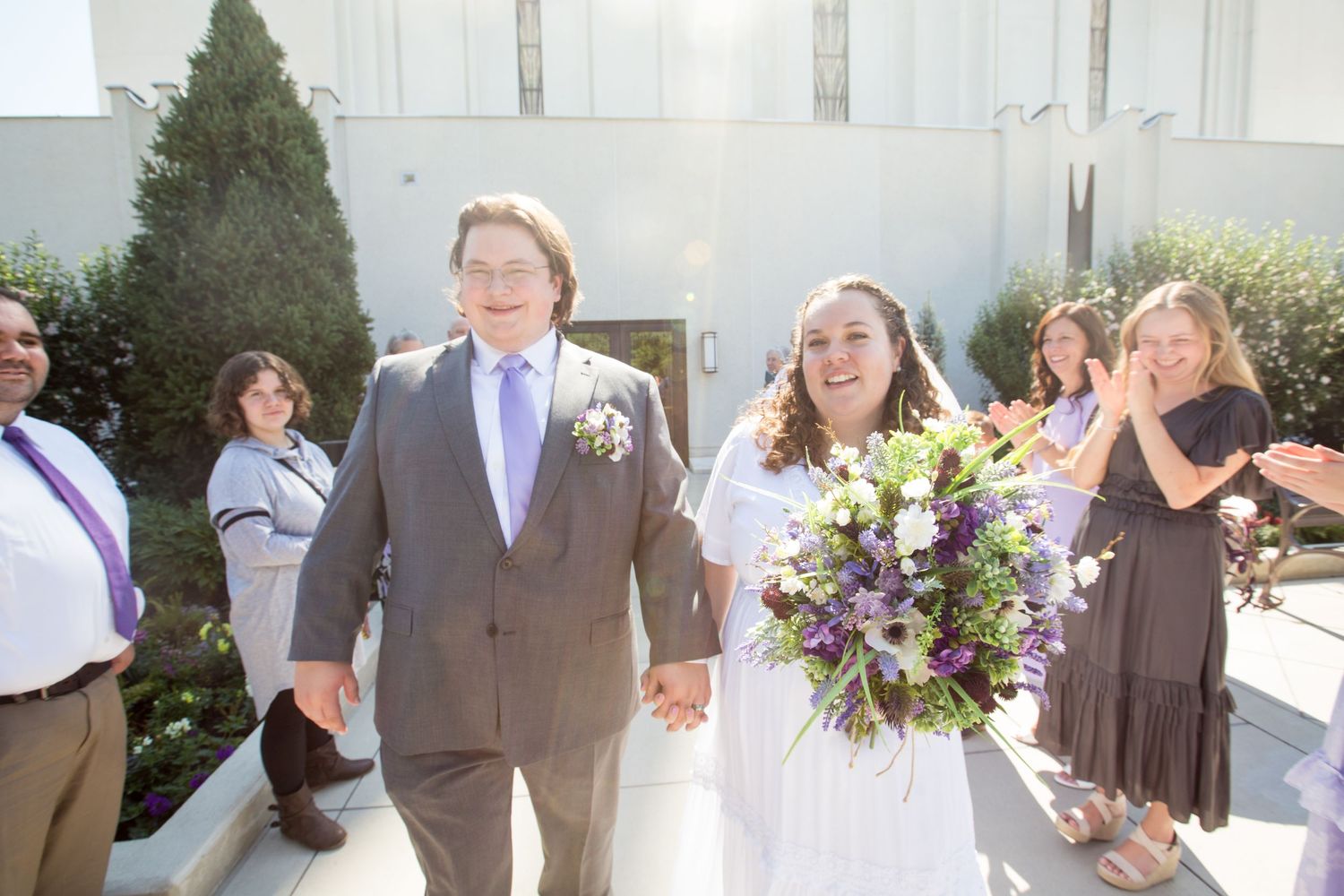 Newlywed couple holding hands and smiling outside a wedding venue, bride holding purple and white bouquet.