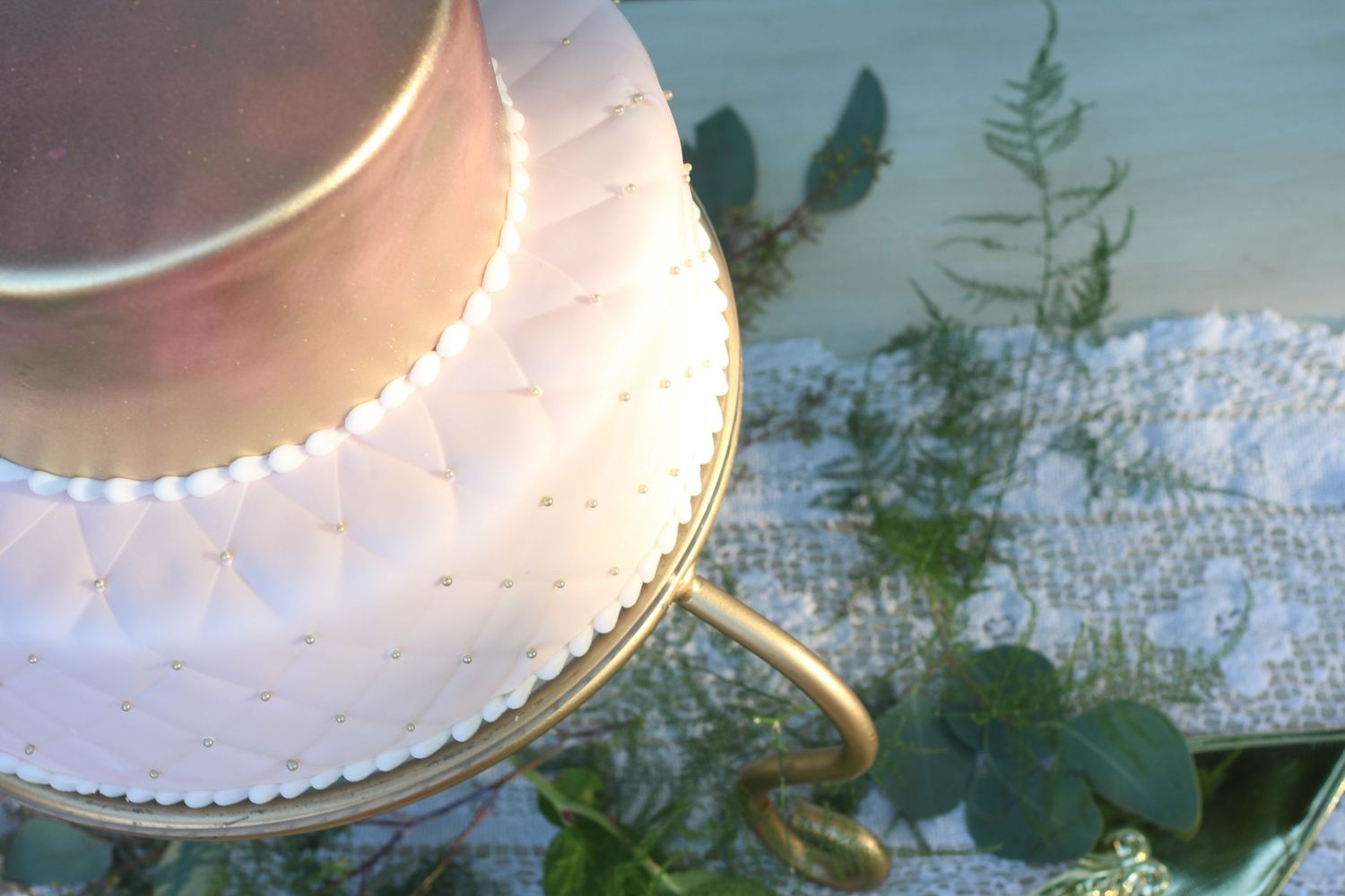 Elegant two-tier white wedding cake with pearl details on a gold stand, surrounded by greenery and lace.