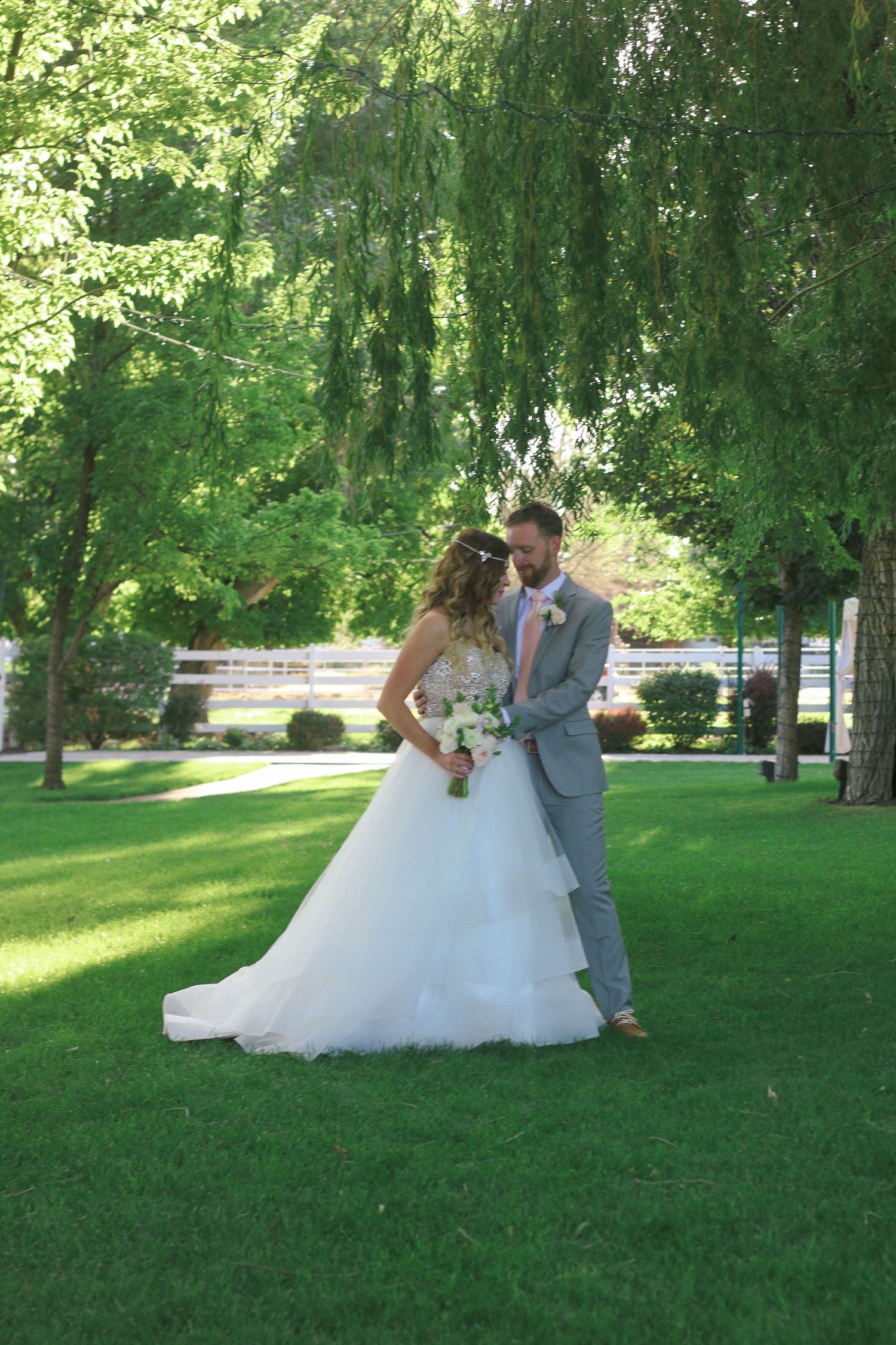 Wedding couple standing on lush green lawn under weeping willow trees.