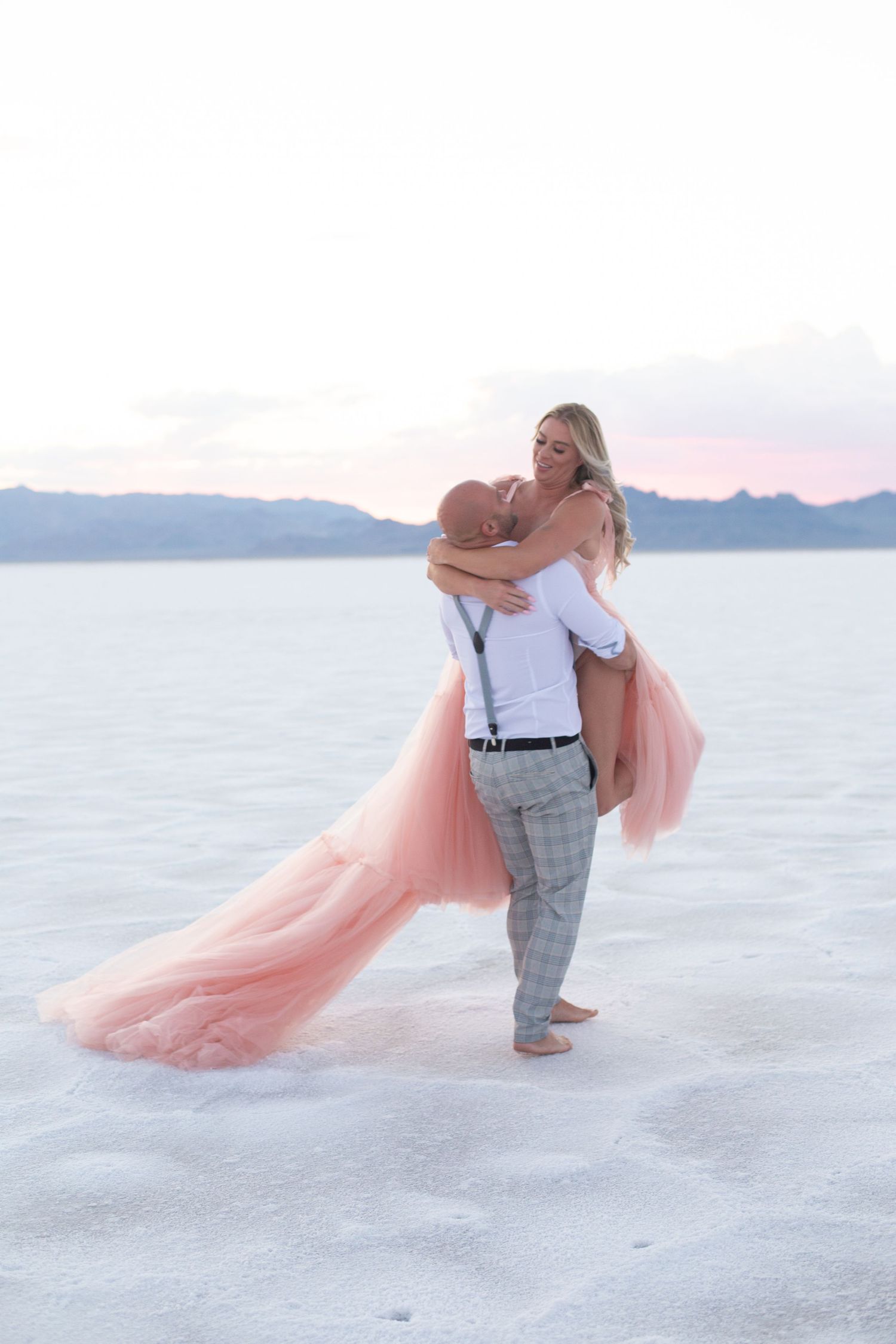 Couple shares intimate moment on white salt flats with pink flowing dress against mountain vista.