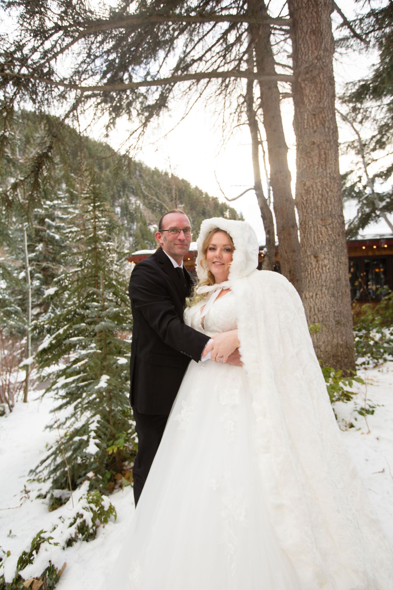 A winter wedding portrait with snow-covered evergreens and a cozy white cape.