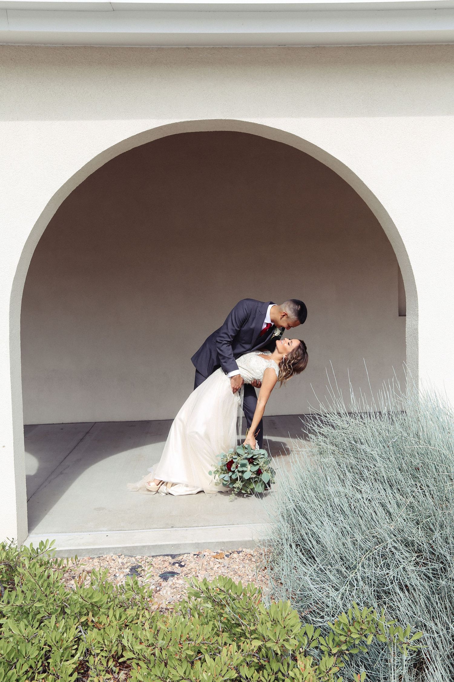A romantic moment captured under arched walkways with decorative landscaping and bushes.