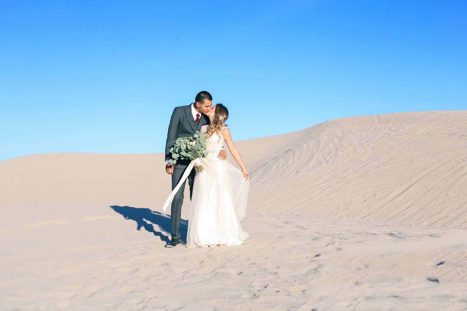 Romantic desert wedding photos with couple posing on sandy dunes against blue sky.
