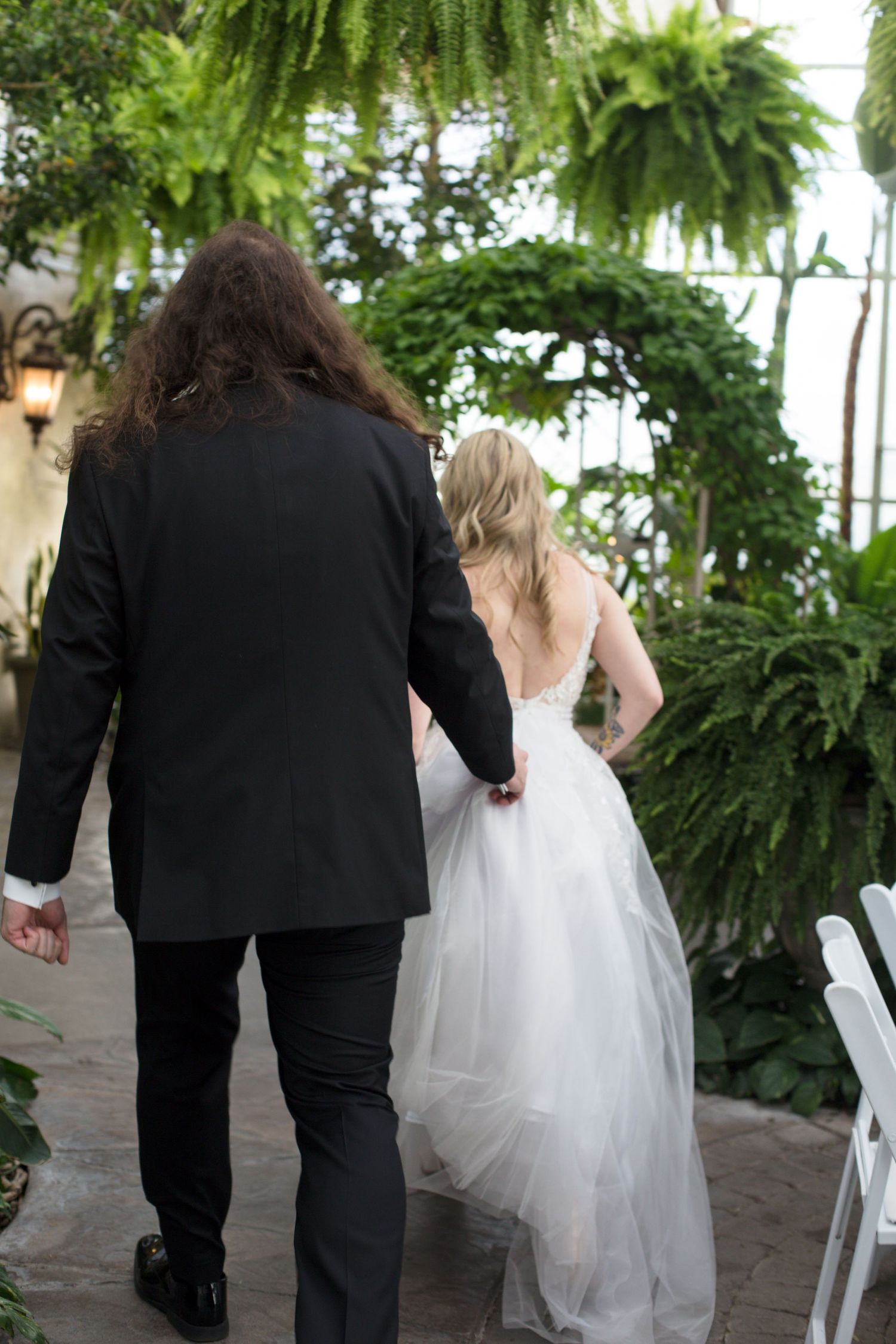 Couple walking through greenhouse venue with lush greenery and white chairs.