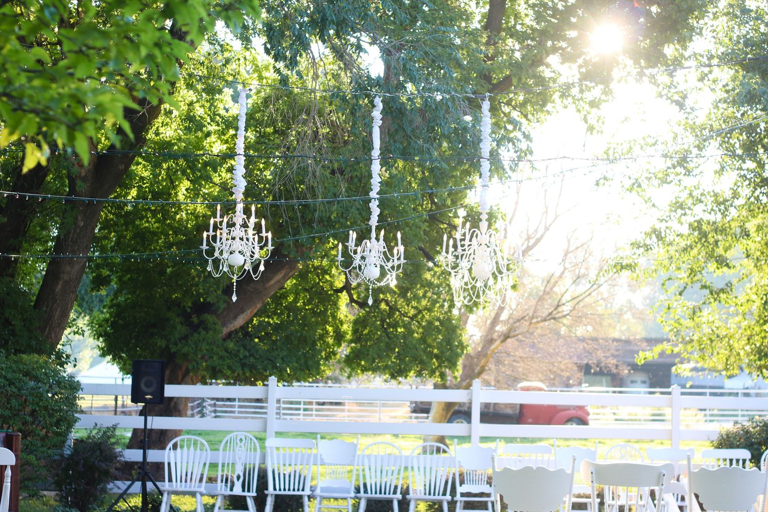 Elegant outdoor wedding venue setup with crystal chandeliers hanging from trees and white chairs at sunset.