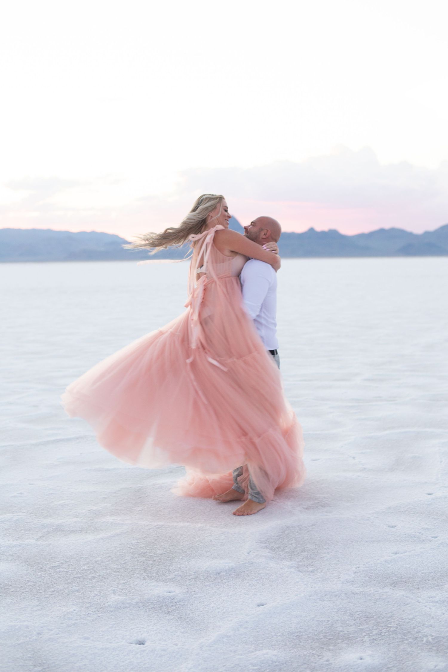 Couple twirls romantically on white salt flats with flowing pink dress at dusk.