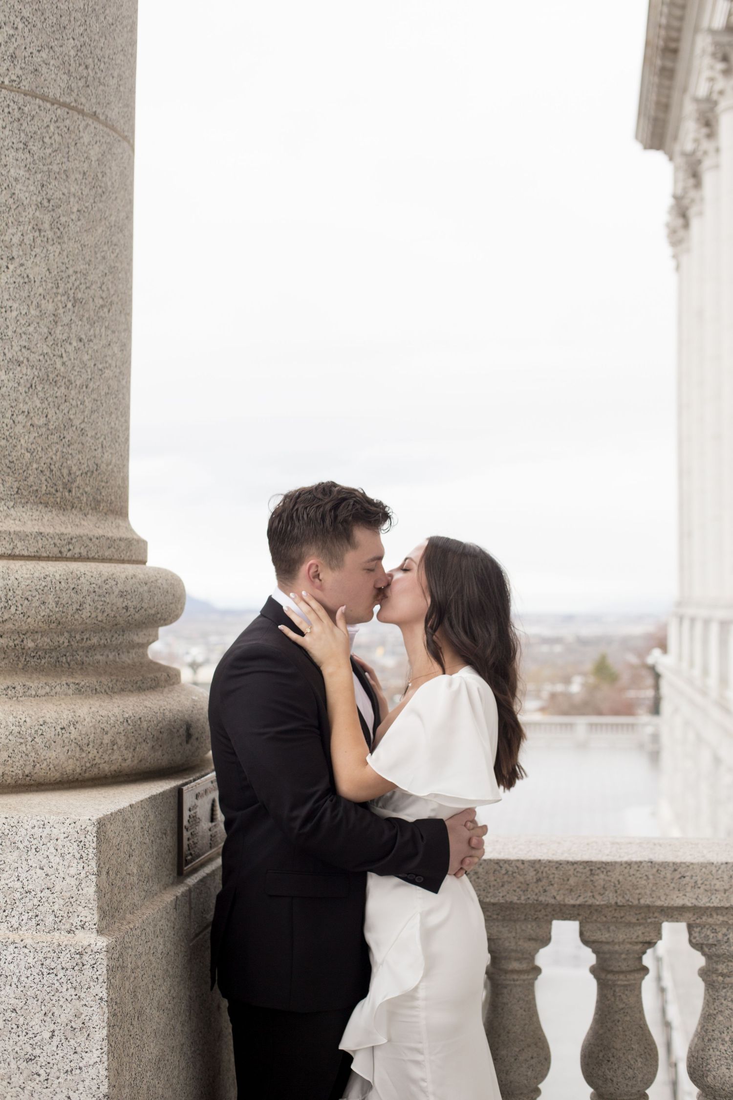 A tender moment between a couple in wedding attire against a stone pillar and balustrade.
