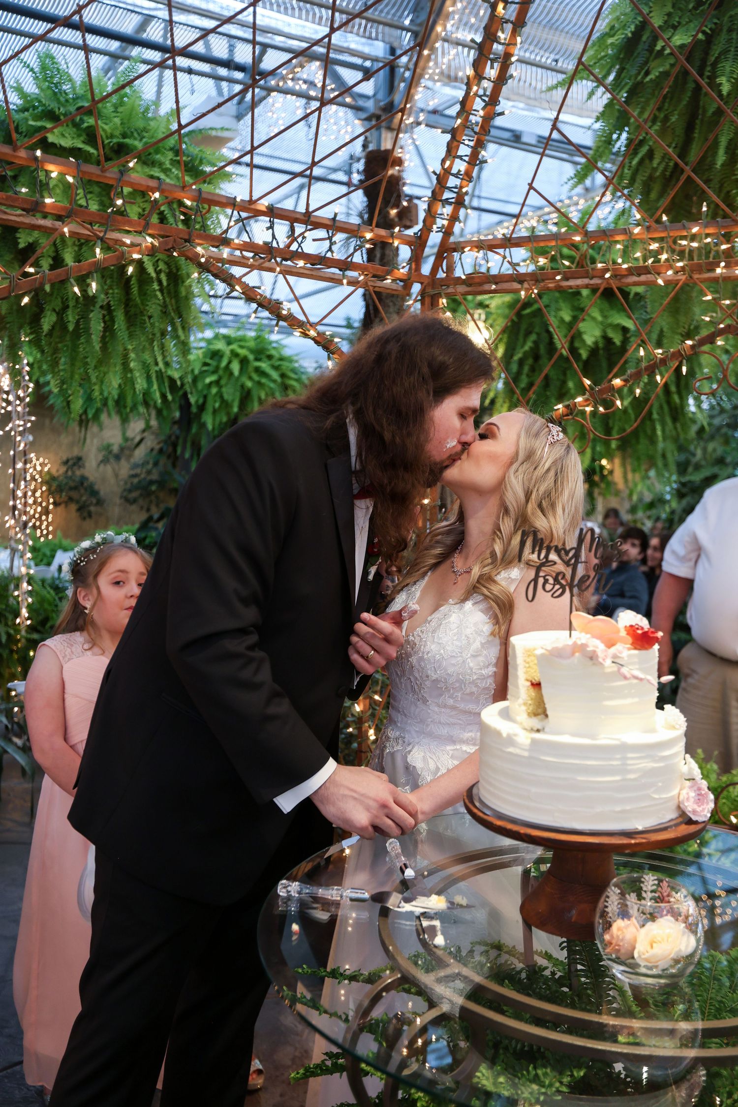 Newlyweds sharing cake cutting moment under twinkling lights in greenhouse venue.