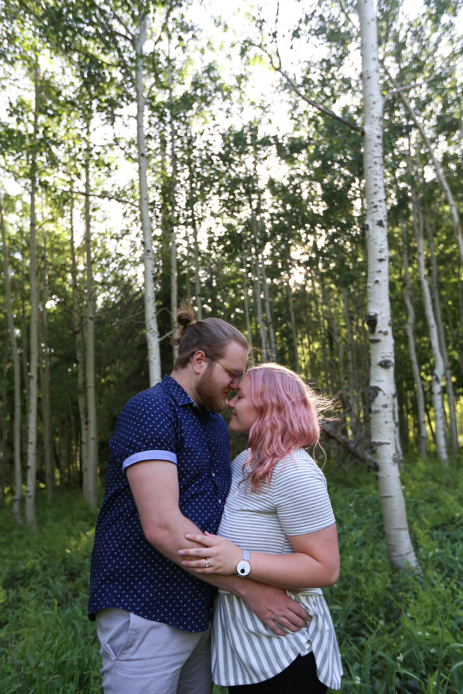 Couple sharing a romantic kiss in a birch tree forest, woman with pink hair embracing bearded man in blue shirt.