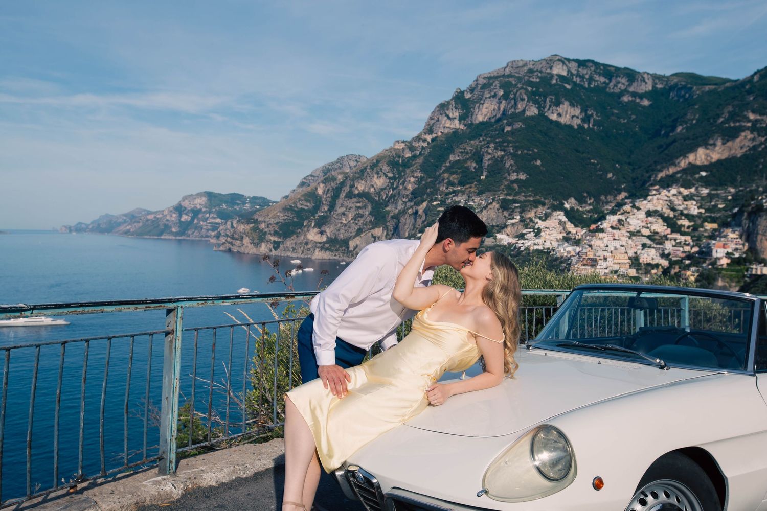 Couple kissing by a vintage white convertible with Amalfi Coast cliffside views in background.