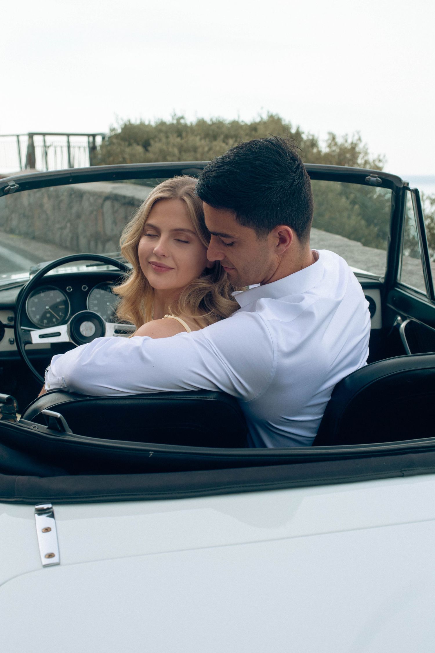 Couple embracing tenderly inside a vintage white convertible with the top down on a scenic overlook.