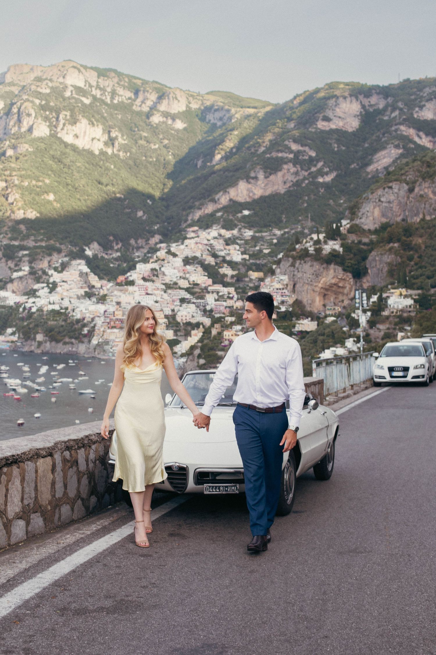 Couple holding hands walking along Amalfi Coast road with white vintage convertible and Positano hillside village behind.