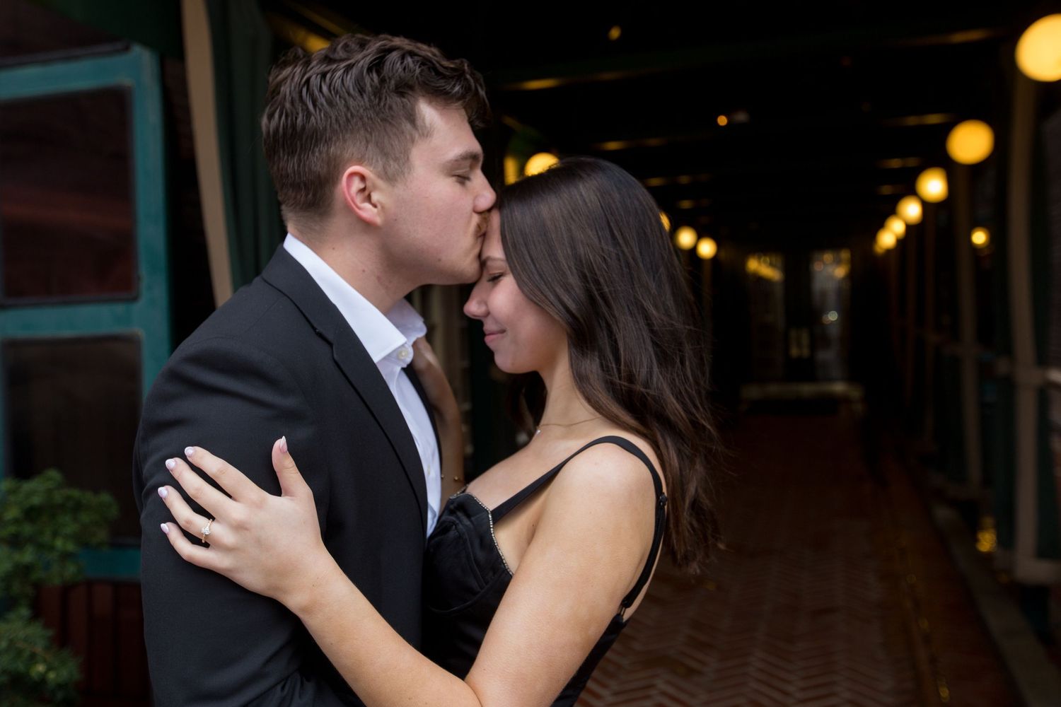 A couple shares a romantic moment, the man kissing the woman's forehead in a warmly lit outdoor corridor.