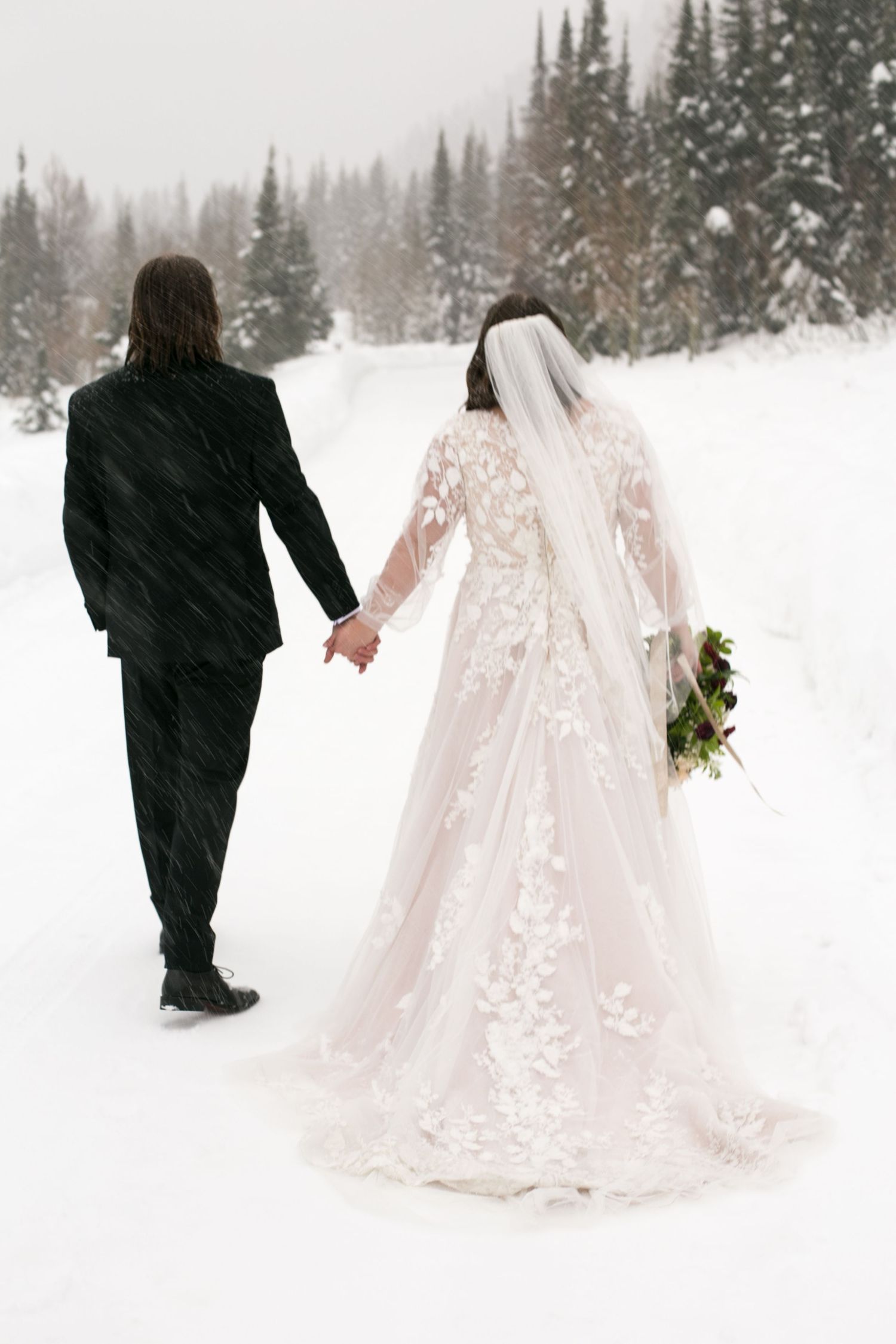 Bride in flowing white dress walks through deep snow with companion.
