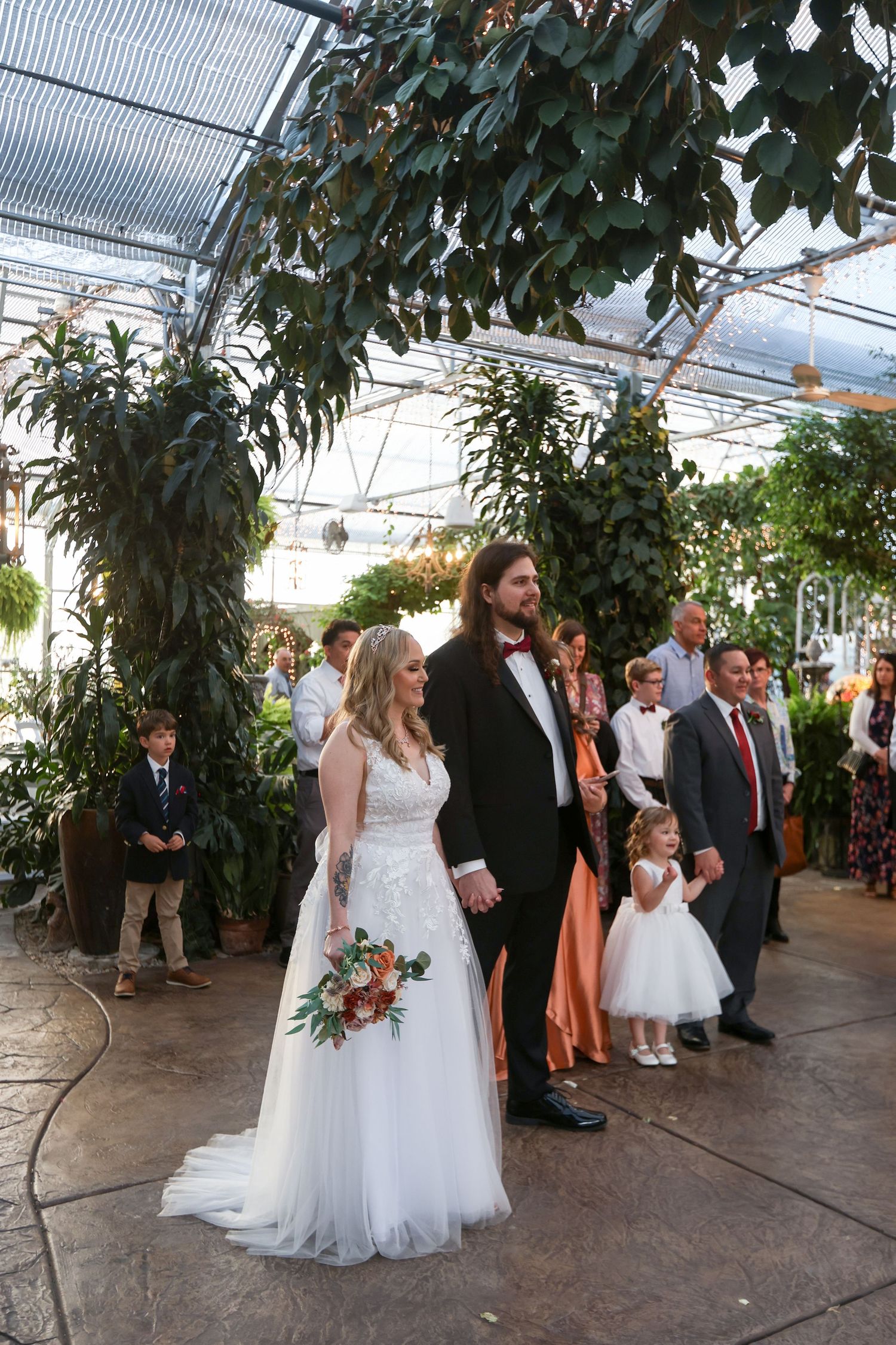 Wedding ceremony taking place in a greenhouse with lush greenery and hanging lights.