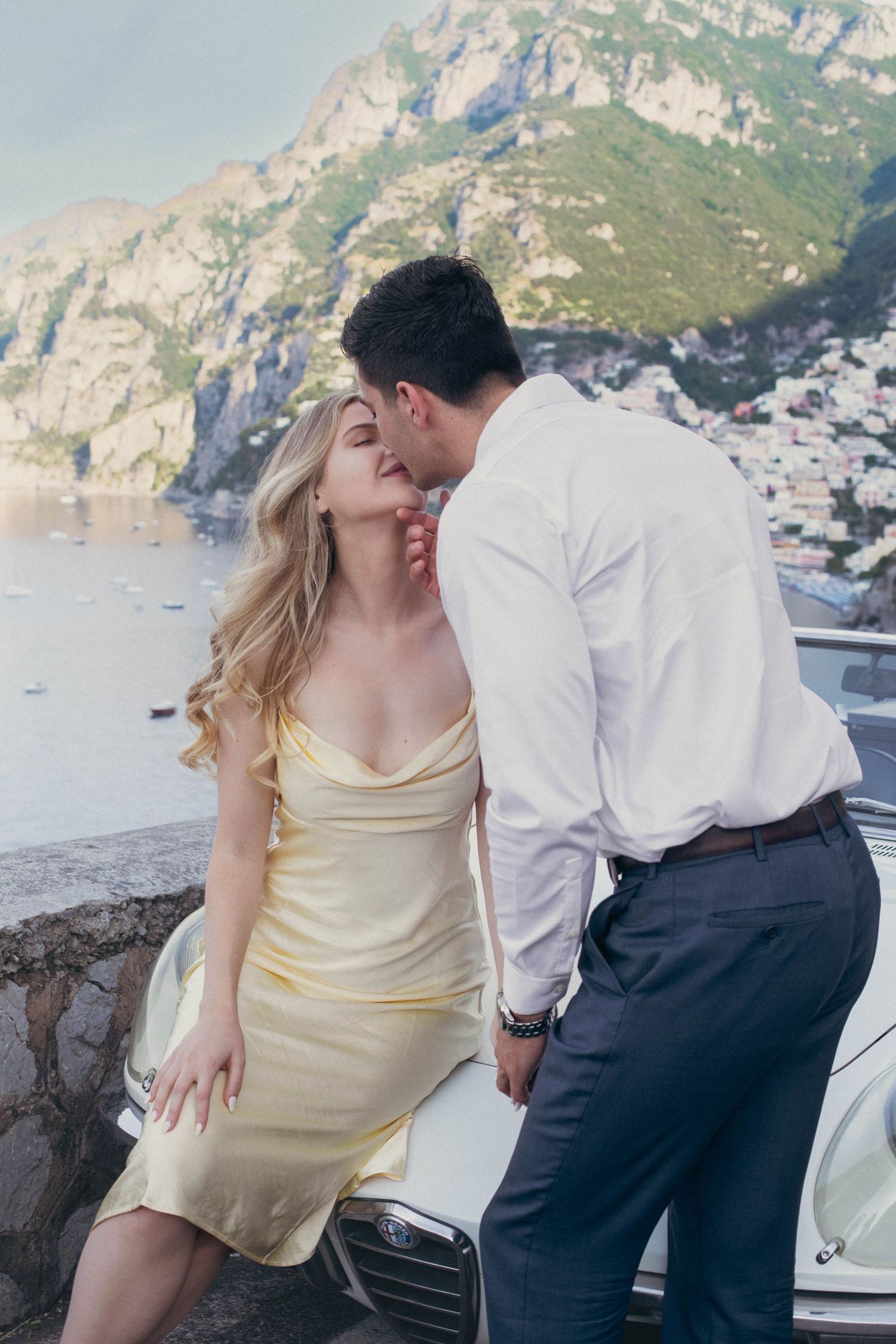 Couple sharing a kiss beside classic white Alfa Romeo convertible on scenic Amalfi Coast road.