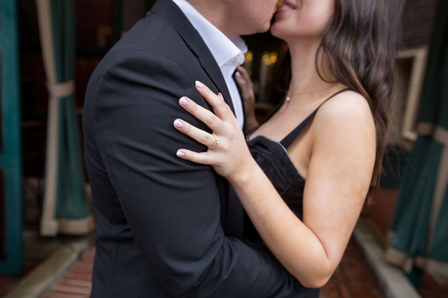 A romantic couple embraces in formal attire against a brick wall background.