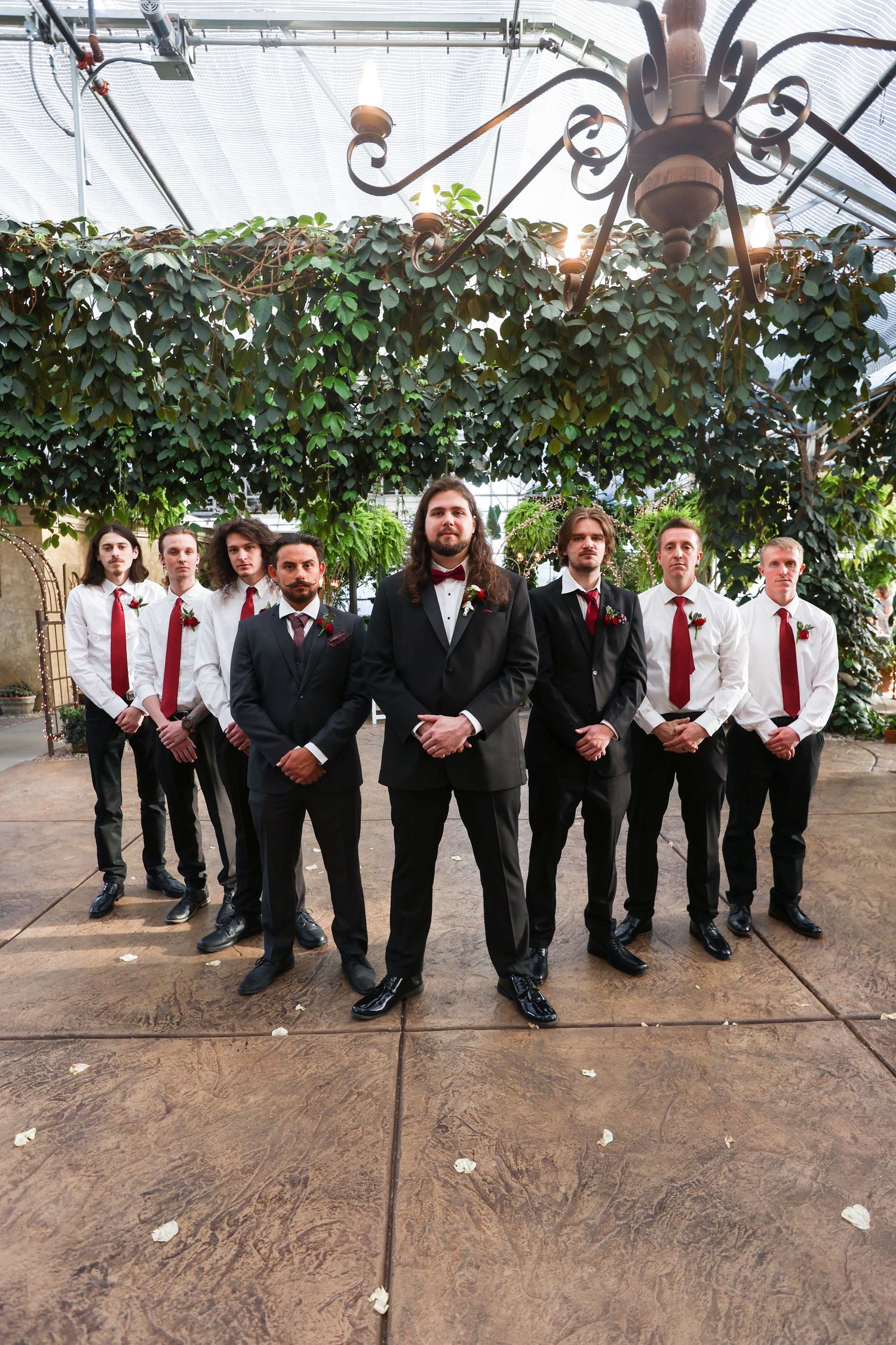 Groomsmen in black suits with red ties stand together under a covered outdoor area.
