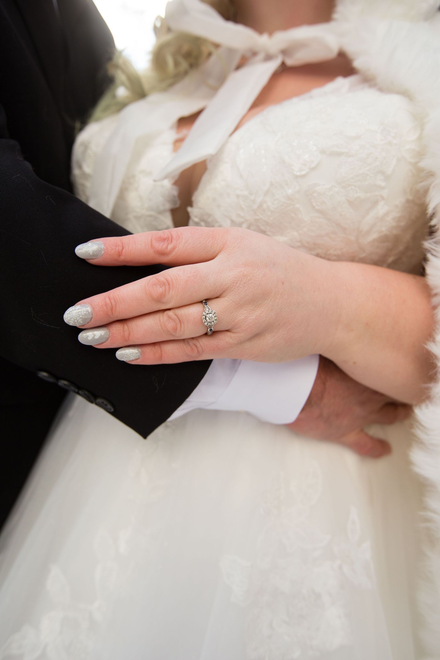 Detail shot of an elegant engagement ring on a bride's manicured hand against white wedding dress fabric.